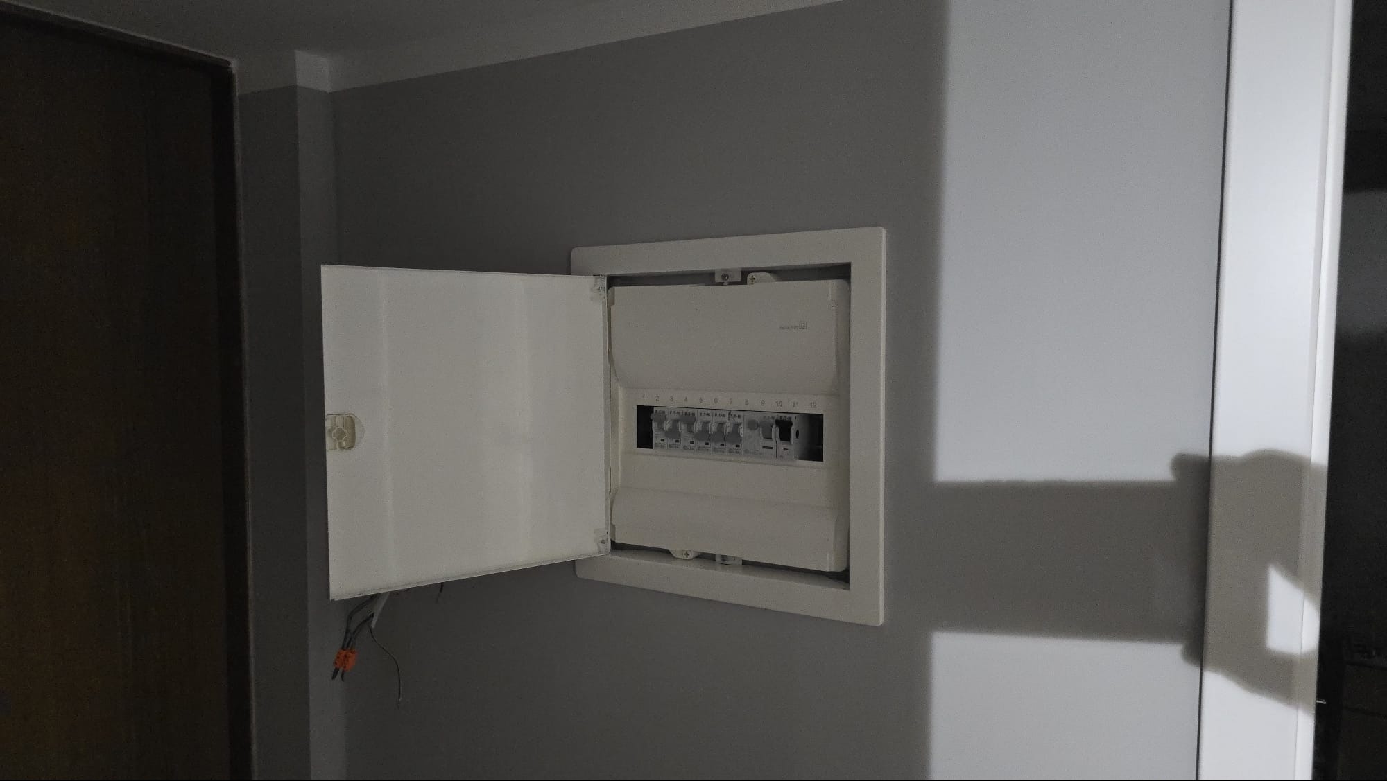 Otwarta skrzynka bezpiecznikowa w ścianie, widoczne przewody. Szara ściana, biała skrzynka z numeracją bezpieczników. Widoczne kable.