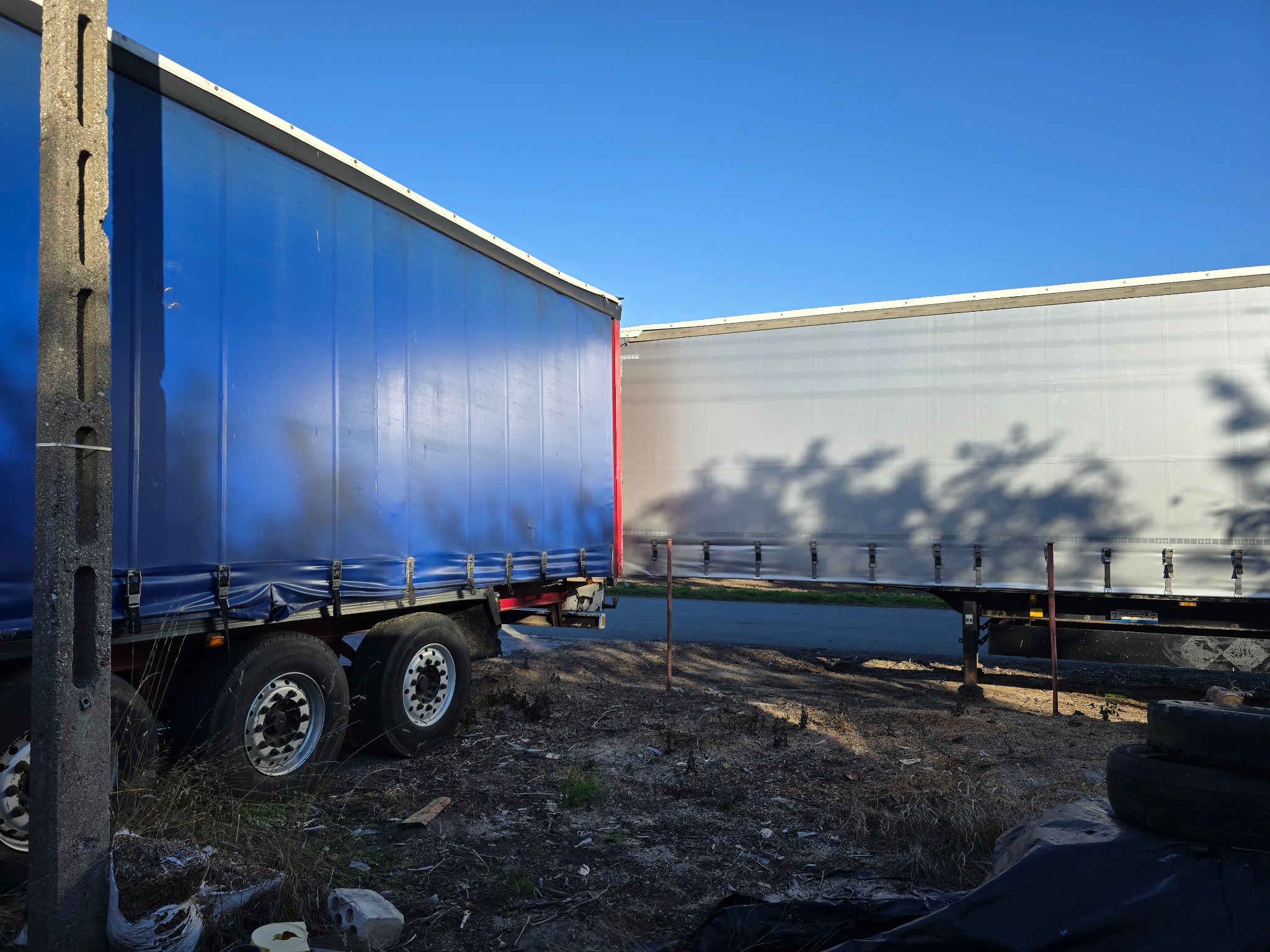 Dwa zaparkowane tiry z naczepami: błękitna i szara, na nieutwardzonym terenie, w tle błękitne niebo. Widoczne opony i fragment słupa betonowego.