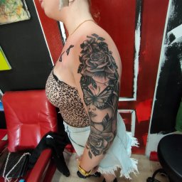 Pracownia Artystyczna Degenera - Kobieta z tatuażem na ramieniu przedstawiającym motywy kwiatowe, motyla i twarz kobiety. Tatuaż w stylu czarno-szarym, z widocznymi detalami. W tle czerwone i czarne elementy dekoracyjne.