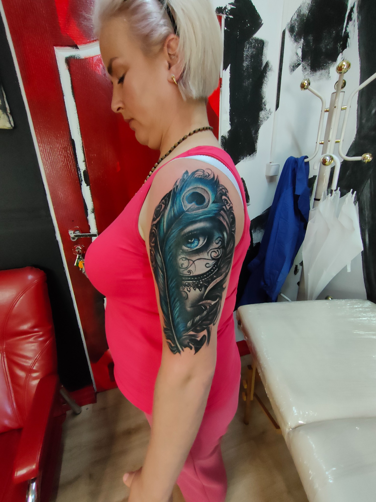 Kobieta z tatuażem na ramieniu przedstawiającym oko i pióro pawie. W tle widoczny fotel w kolorze czerwonym i białe łóżko do wykonywania tatuaży.
