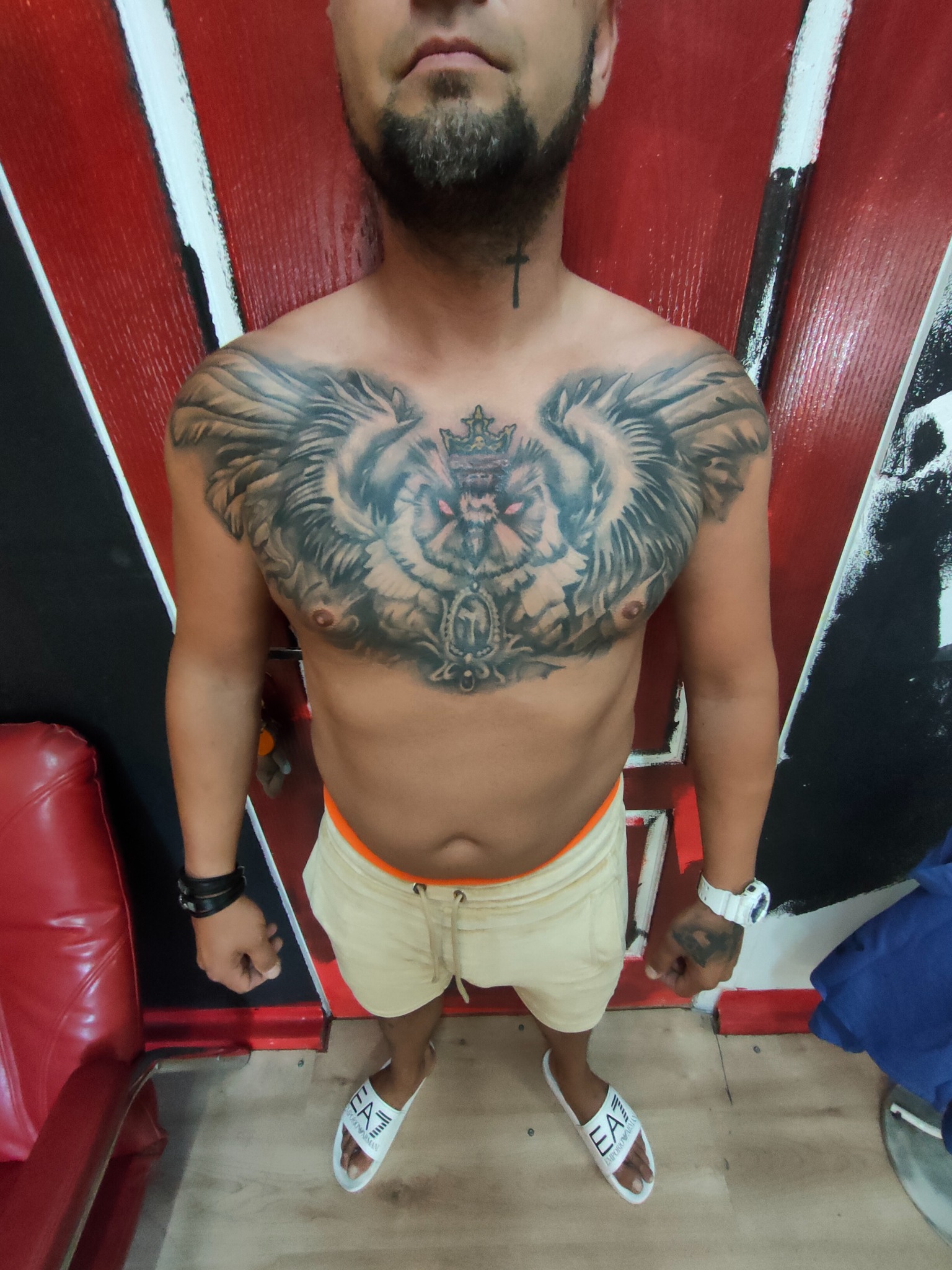 Mężczyzna z dużym tatuażem orła na klatce piersiowej i tatuażem krzyża na szyi, stojący na tle ściany z czerwono-czarnymi akcentami.