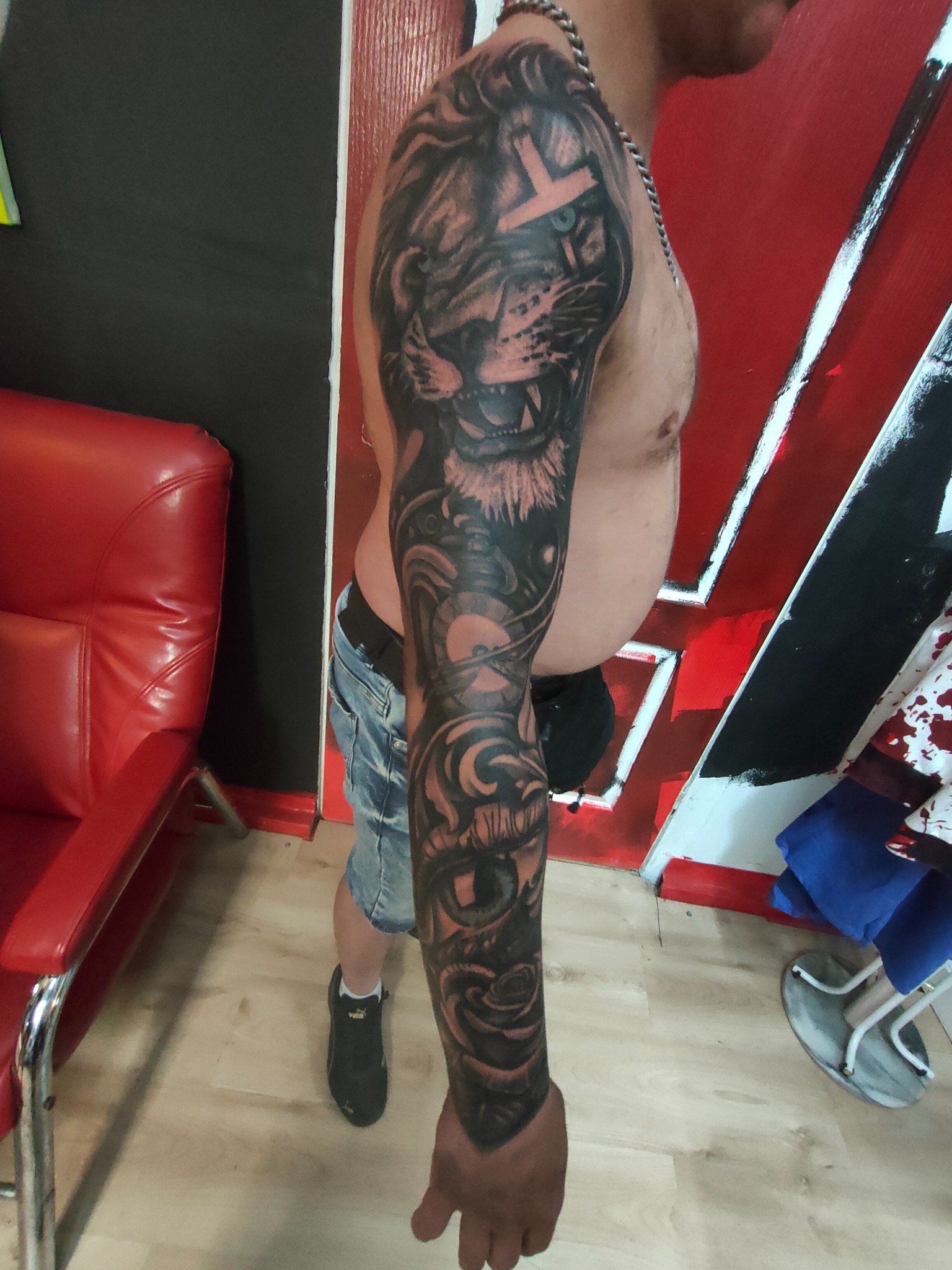 Męskie ramię z czarno-szarym tatuażem: lew z symbolem na czole, oko i róża. W tle czerwono-biała ściana i czerwone krzesło. Widoczne fragmenty ciała i ubrania.