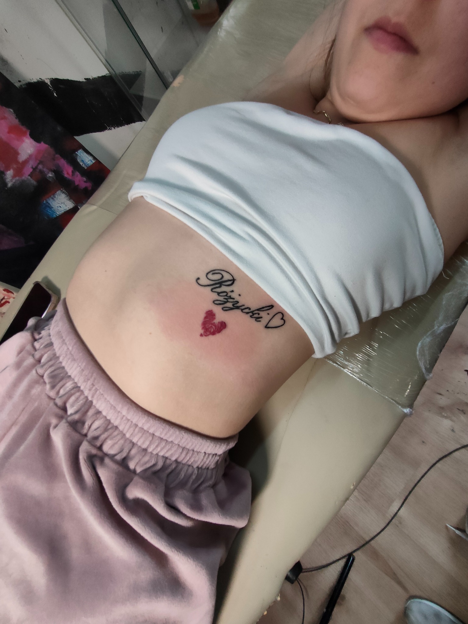 Kobieta leży na leżance z nowym tatuażem na brzuchu: napis kursywą 'Różycki' z sercem. Skóra zaczerwieniona wokół tatuażu.