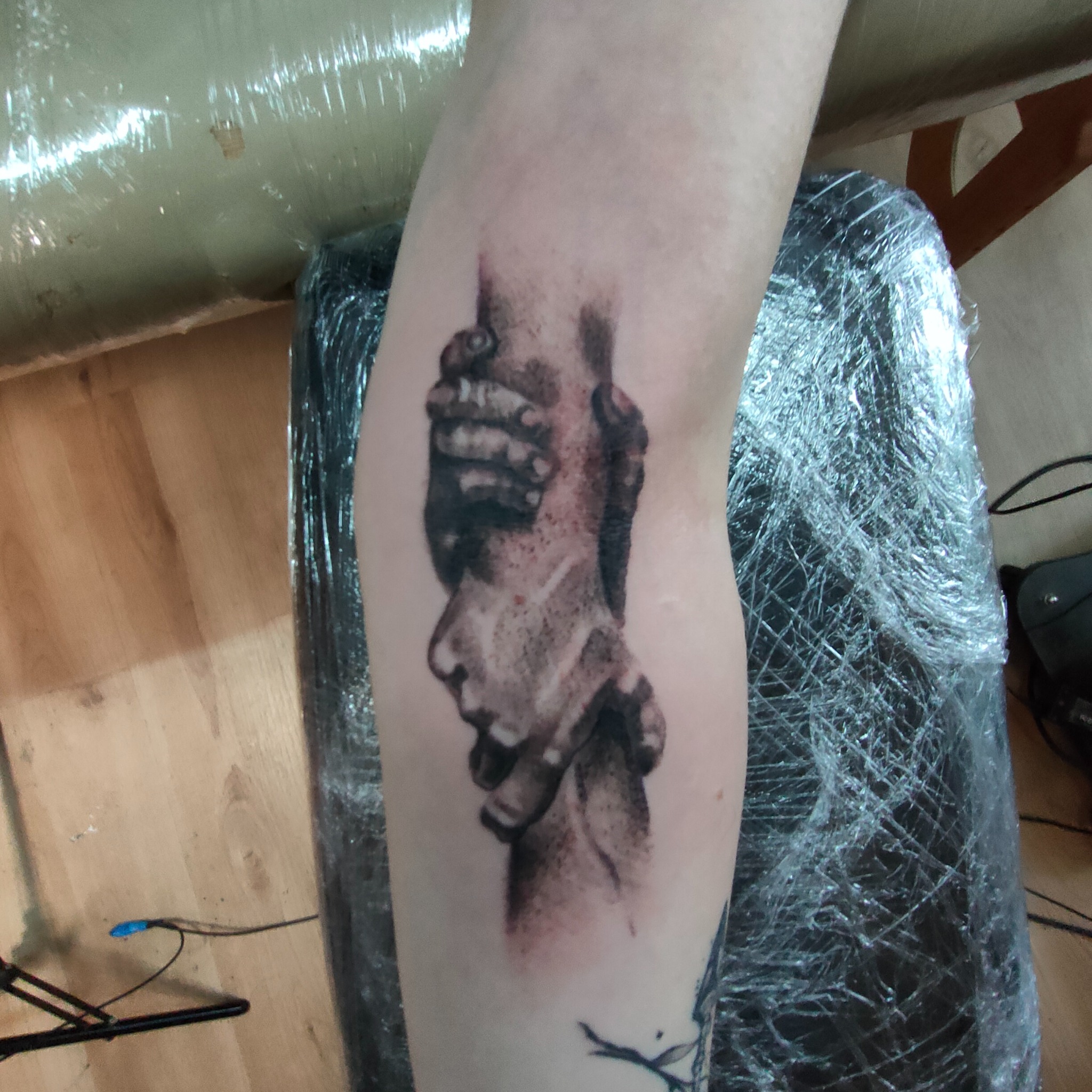 Czarno-biały tatuaż przedstawiający splecione dłonie na przedramieniu, świeżo po wykonaniu, zabezpieczony folią ochronną. Widoczne detale cieniowania i konturów.