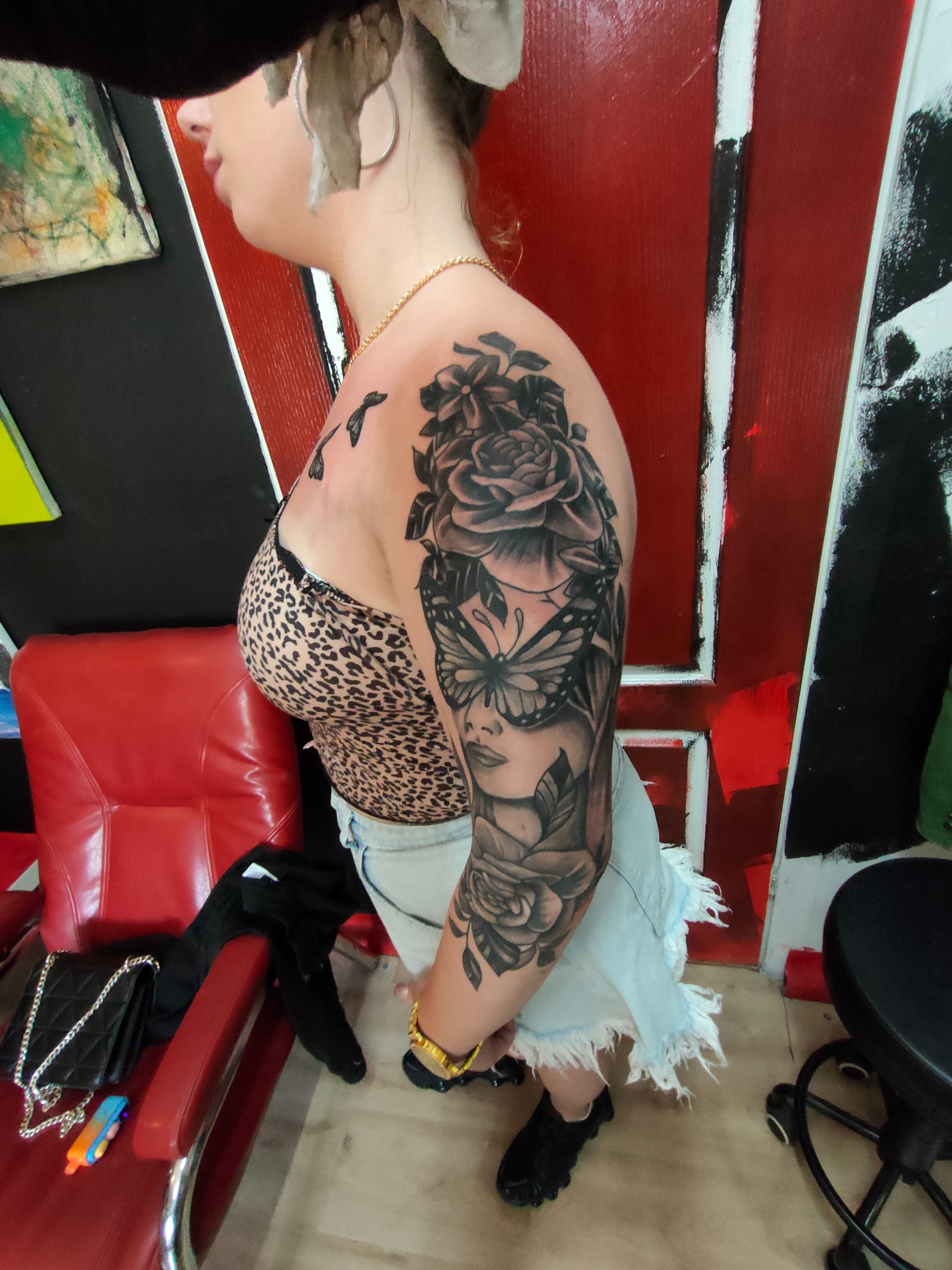 Kobieta z tatuażem na ramieniu przedstawiającym motywy kwiatowe, motyla i twarz kobiety. Tatuaż w stylu czarno-szarym, z widocznymi detalami. W tle czerwone i czarne elementy dekoracyjne.