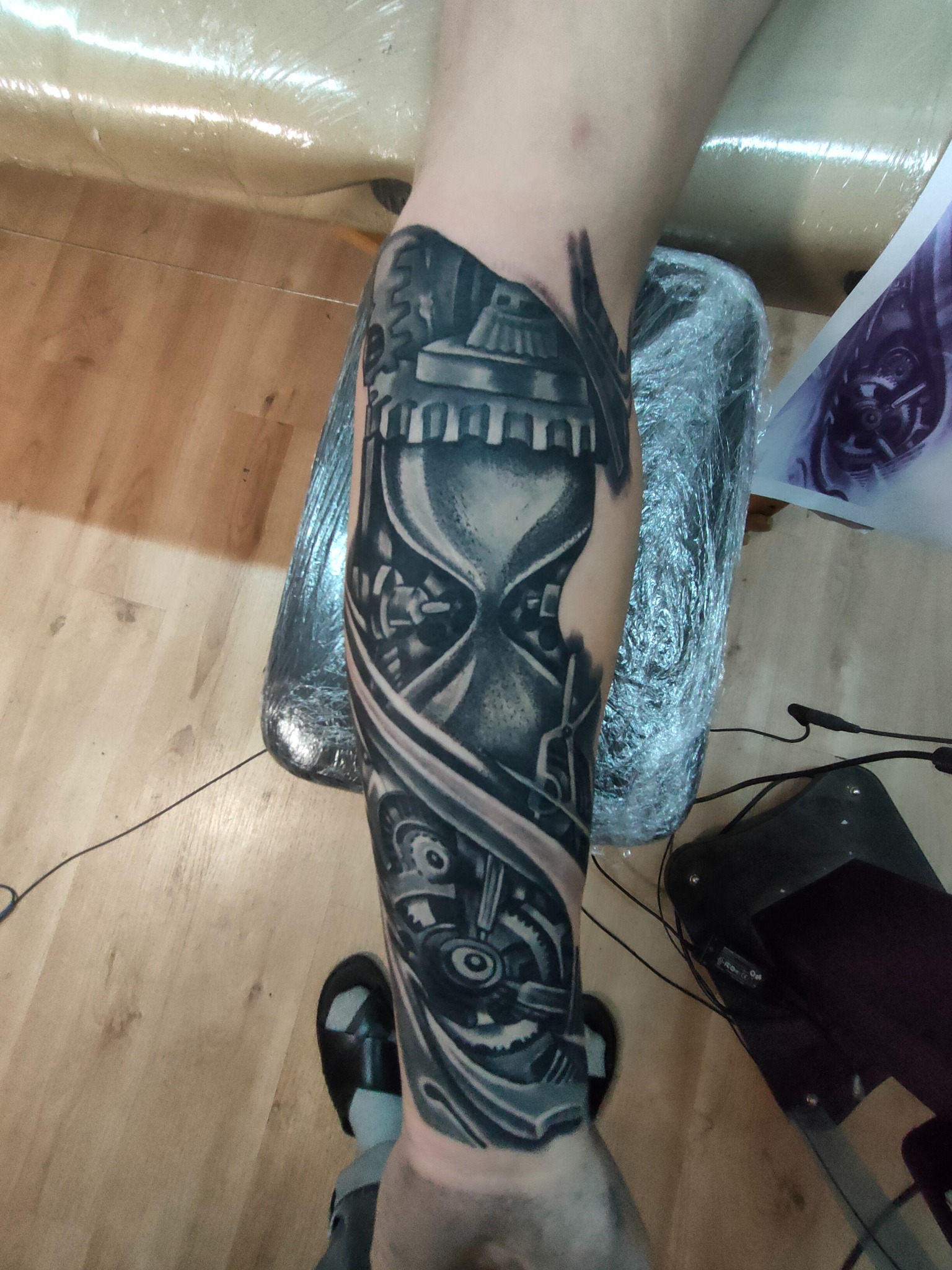 Czarno-szary tatuaż na przedramieniu przedstawiający klepsydrę w stylu steampunk, otoczoną mechanizmami i zębatkami. Widoczne detale, świeżo po wykonaniu, zabezpieczony folią.