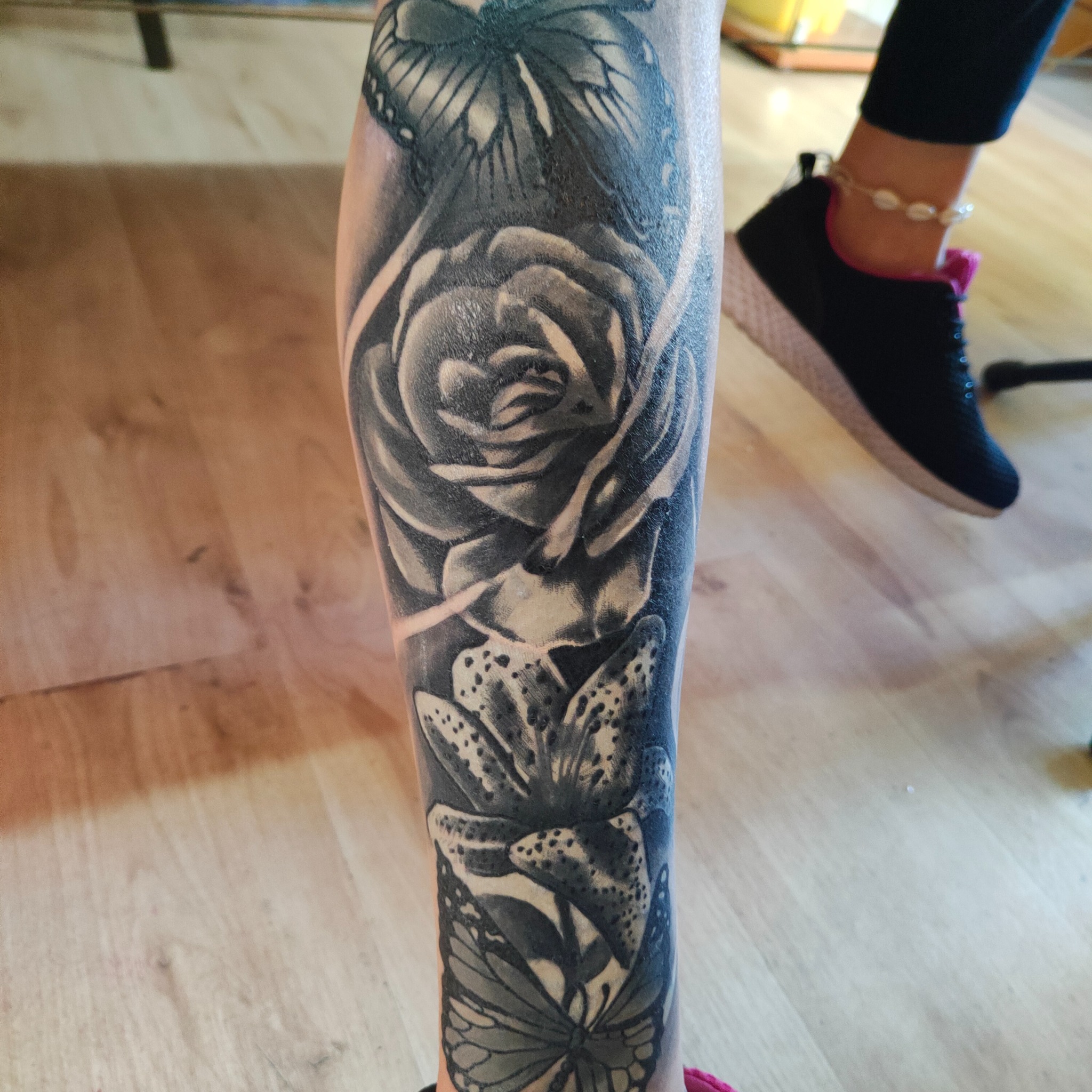 Czarno-biały tatuaż na łydce przedstawiający motyle, różę i lilię. Widoczne fragmenty buta i podłogi. Realistyczny styl tatuażu.