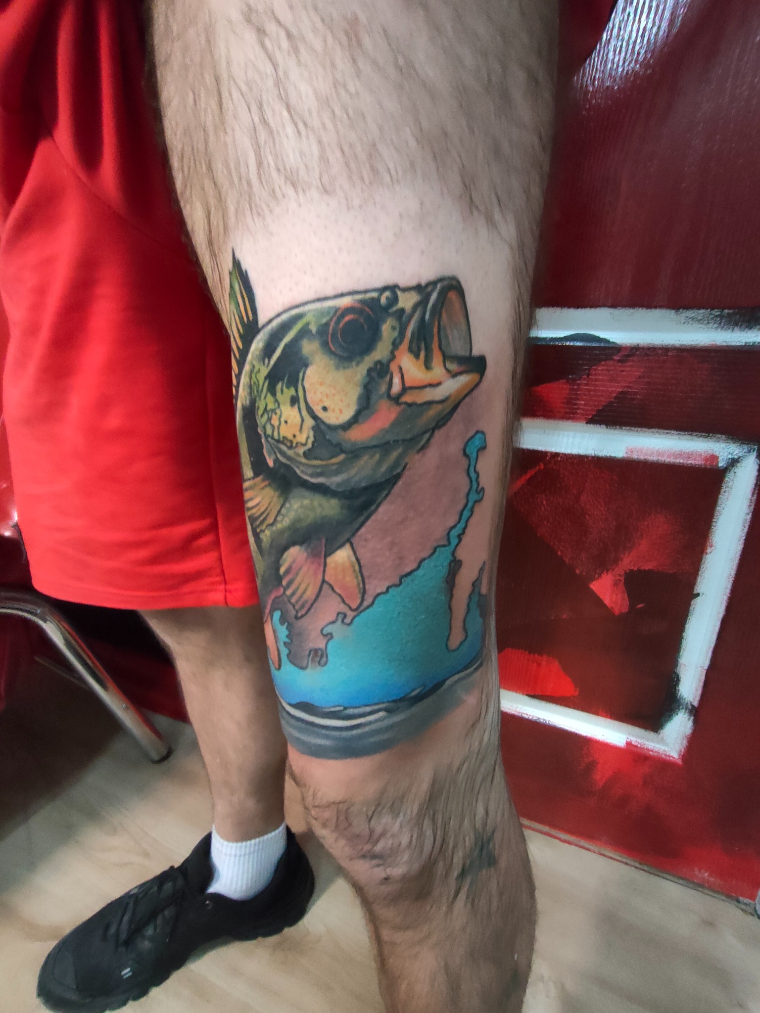 Tatuaż okonia na nodze mężczyzny, z błękitnym tłem imitującym wodę. Widoczna stopa w czarnym bucie i fragment czerwonej ściany z białym wzorem.