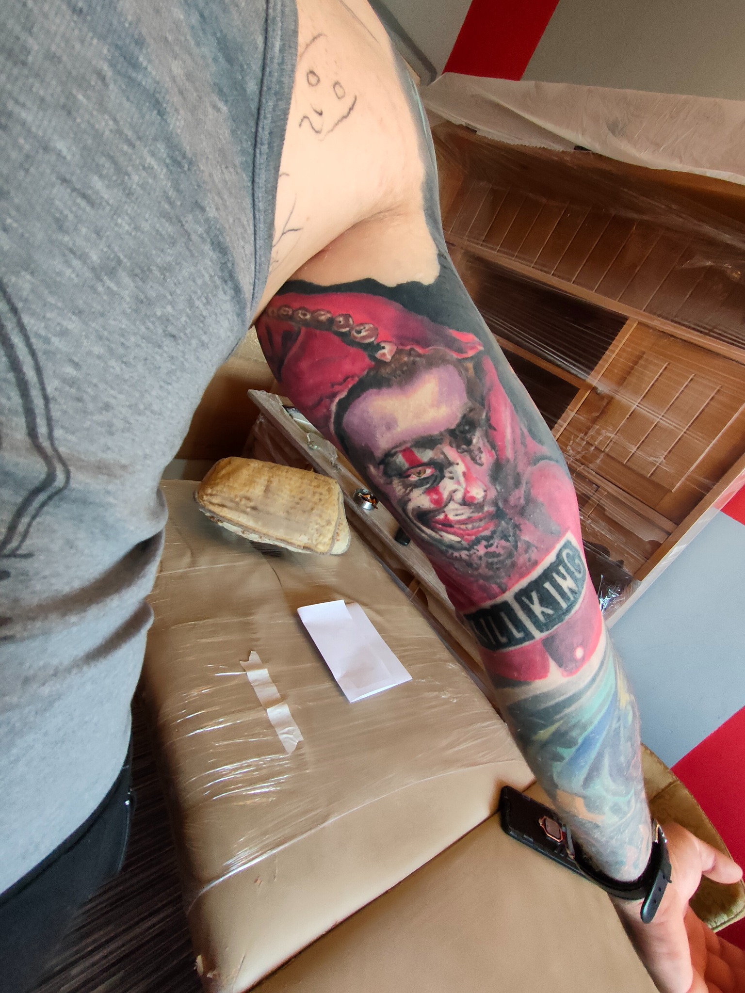 Ramię z kolorowym tatuażem przedstawiającym postać z filmu 'The Shining', leżące na powierzchni z telefonem i kartką papieru w pobliżu owiniętego mebla.