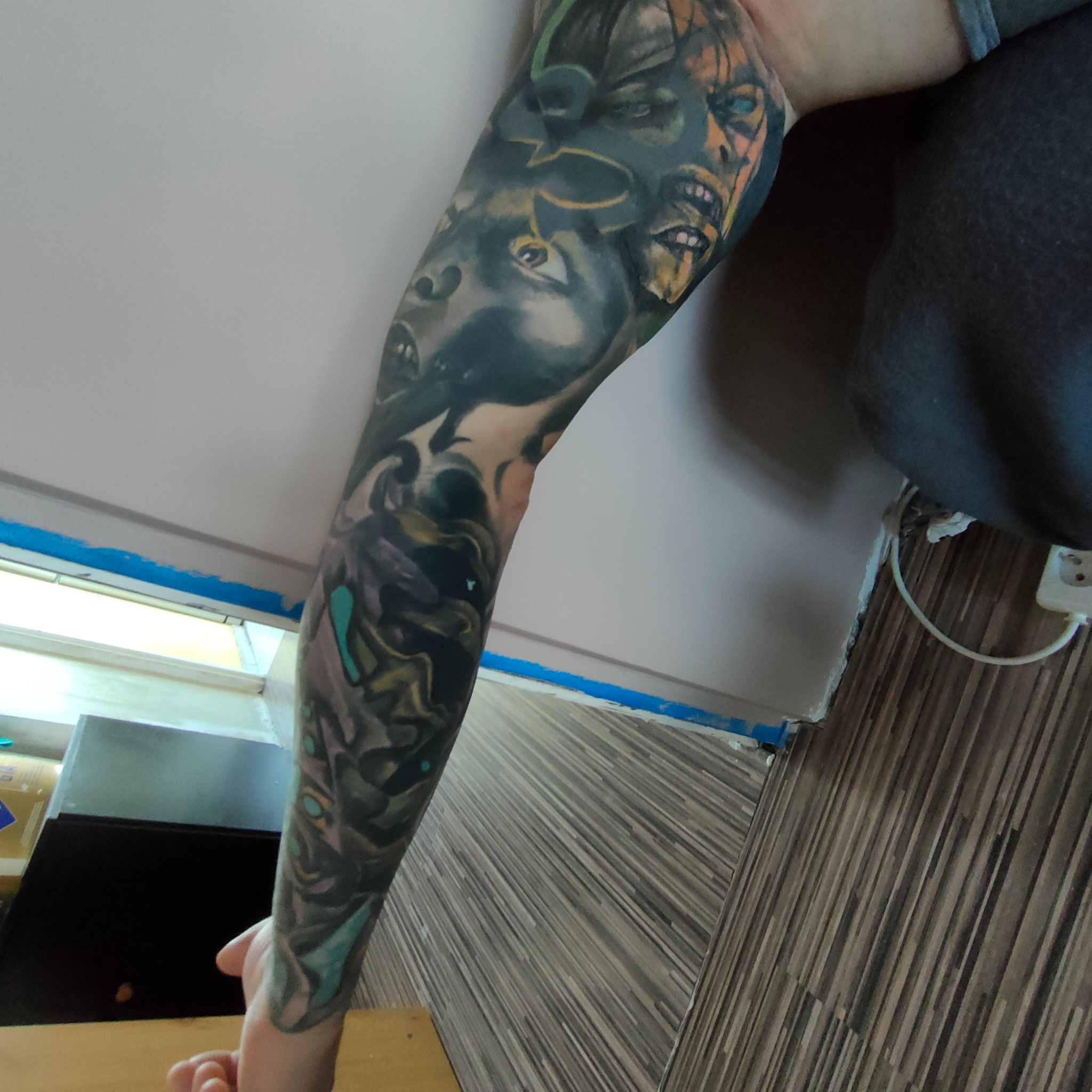 Ręka z tatuażem przedstawiającym fantastyczne postacie, z detalami cieniowania i kolorowymi akcentami. Widoczne fragmenty otoczenia, w tym ściana i podłoga.