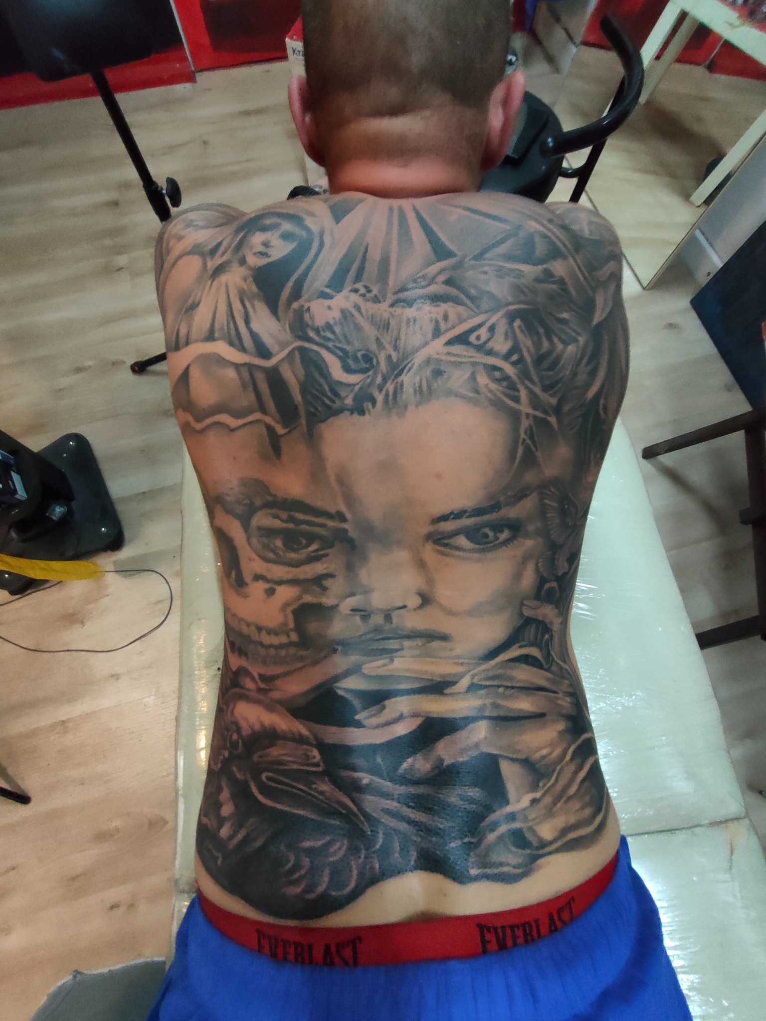 Czarno-biały, szczegółowy tatuaż na plecach mężczyzny przedstawiający portret kobiety, czaszkę i kruka. Widok z tyłu, skupienie na artystycznym wzorze.