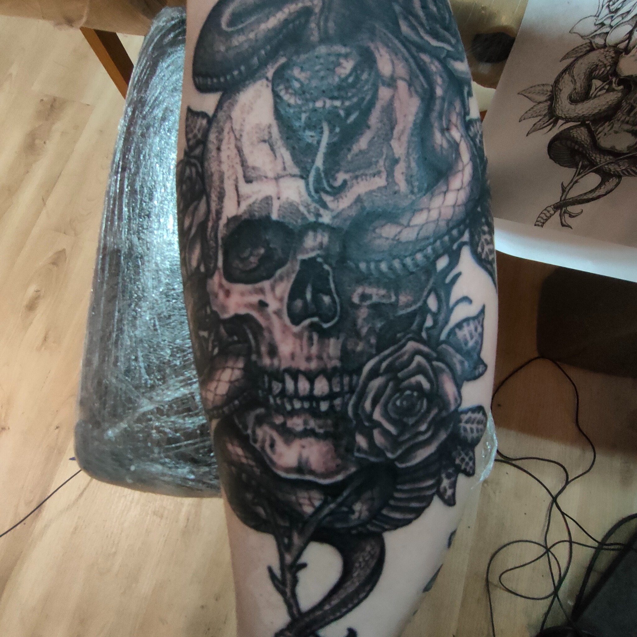 Czarno-biały tatuaż na ramieniu przedstawiający czaszkę oplecioną wężem i różą, zabezpieczony folią ochronną. Obok widoczny szkic podobnego wzoru.