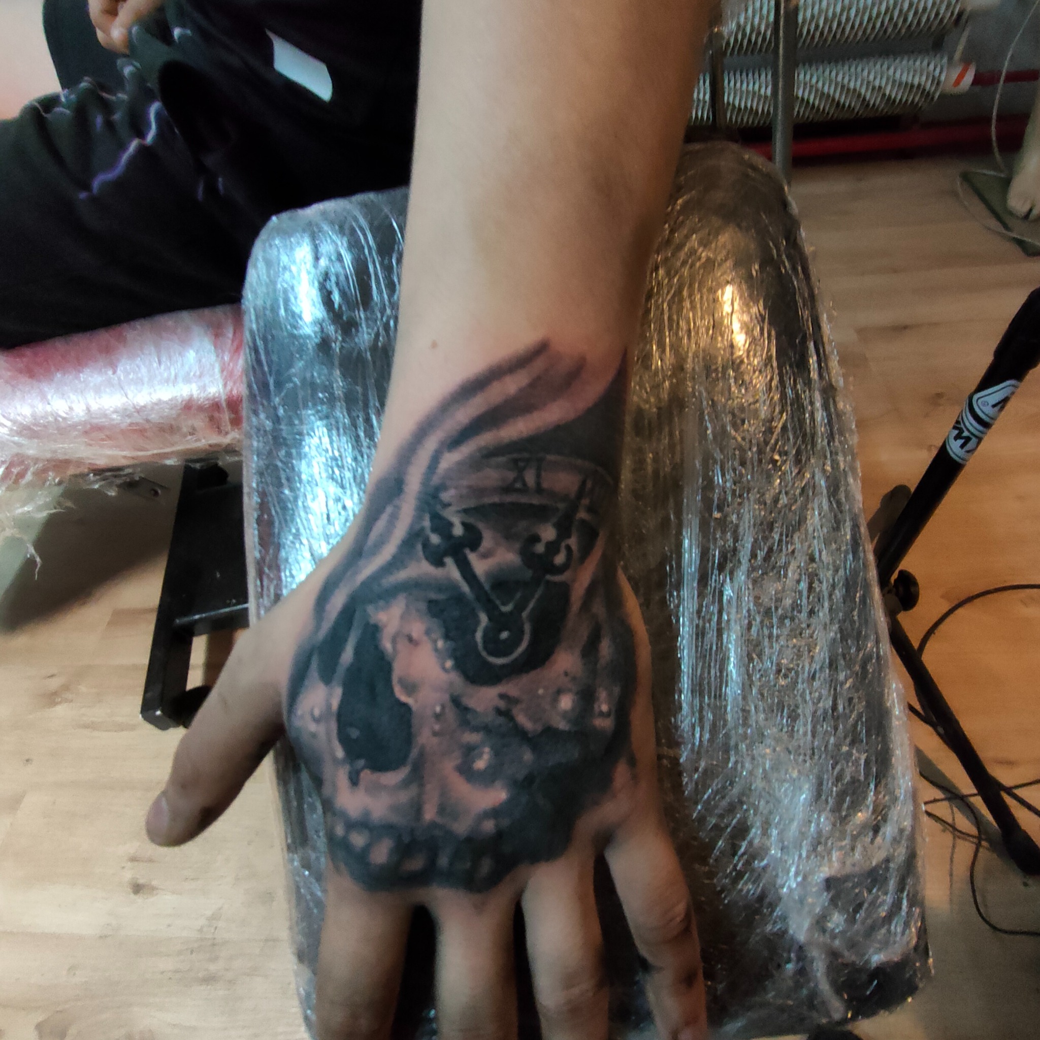 Czarno-szary tatuaż na dłoni przedstawiający czaszkę i zegar, wykonany w studiu tatuażu. Widoczne detale cieniowania i konturów.