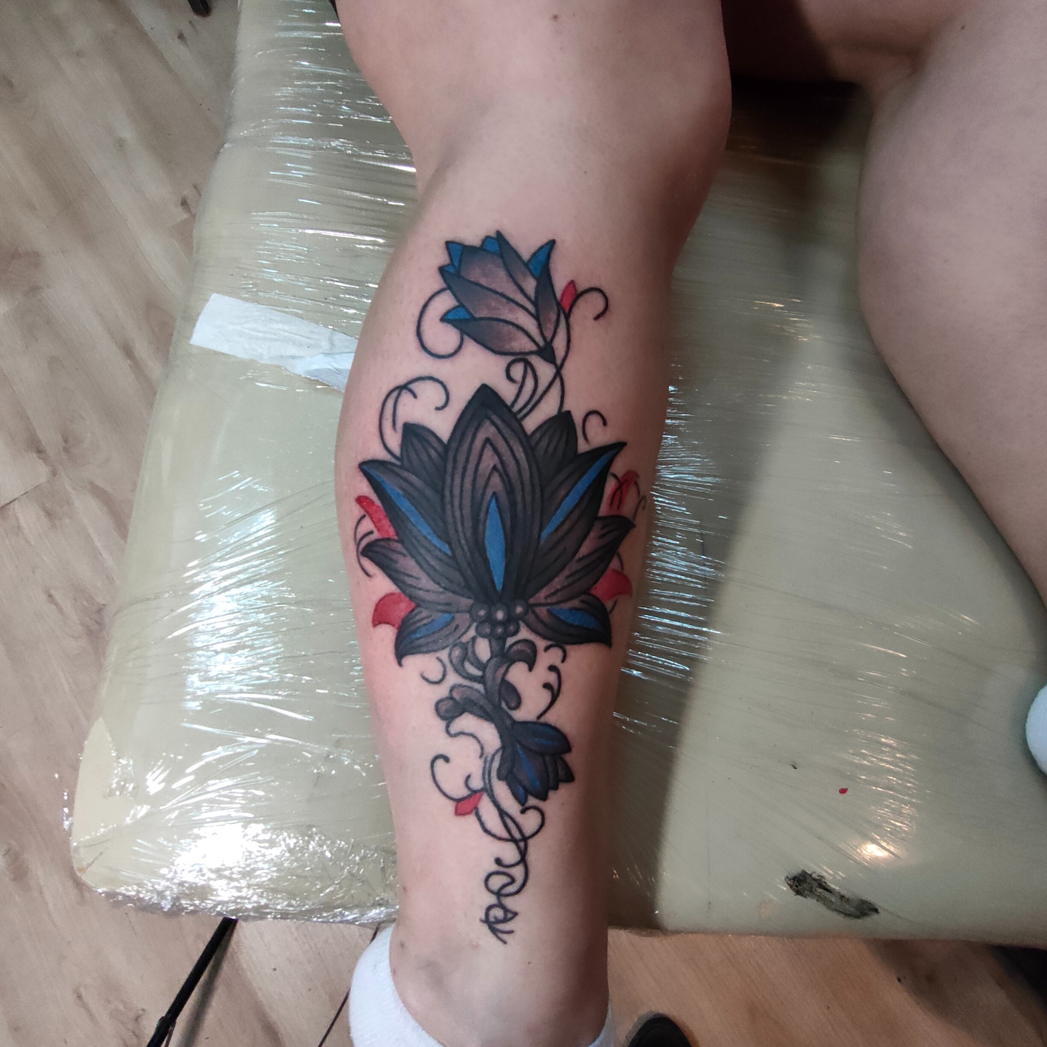 Świeży tatuaż na łydce z motywem kwiatowym w kolorach szarości, czerni, błękitu i czerwieni. Skóra wokół tatuażu zaczerwieniona, zabezpieczona folią. Widoczny fragment drewnianej podłogi.