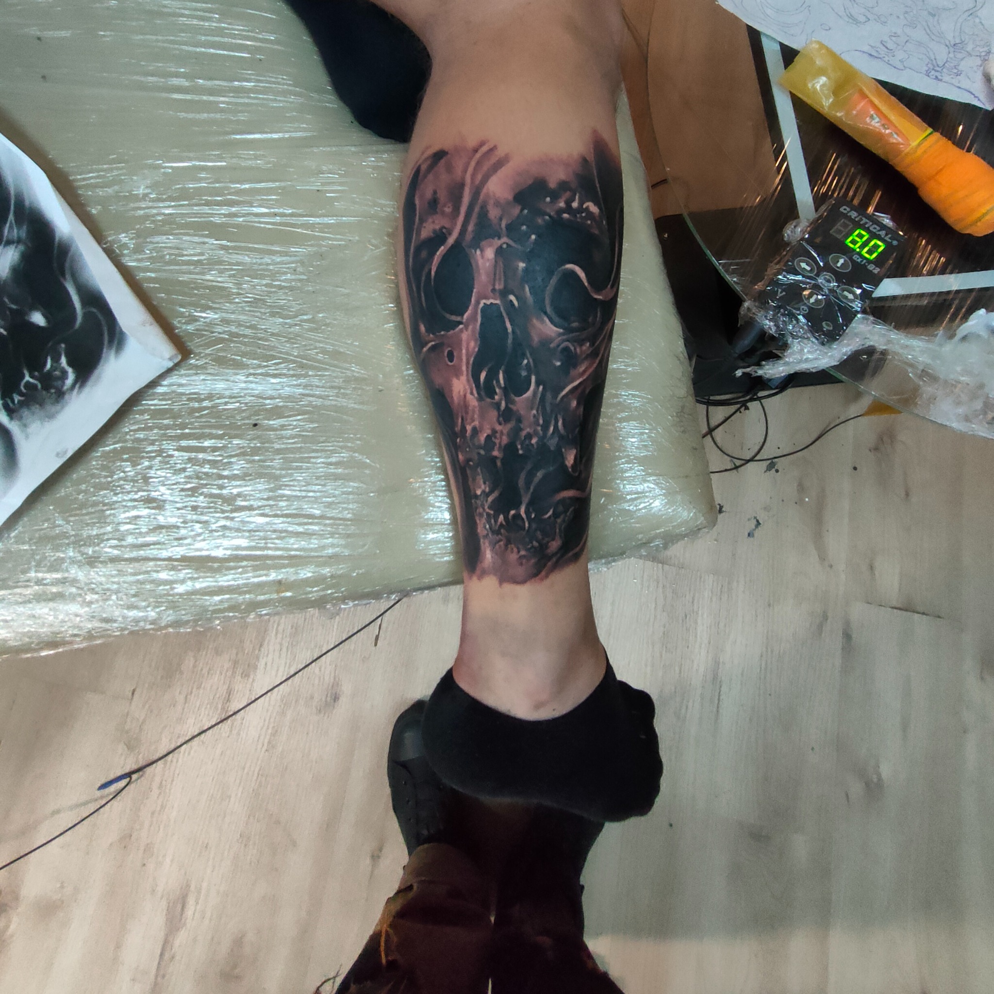 Tatuaż czaszki na łydce, wykonany w technice blackwork. Widoczna maszyna do tatuażu i kalka z wzorem. Noga owinięta folią ochronną.