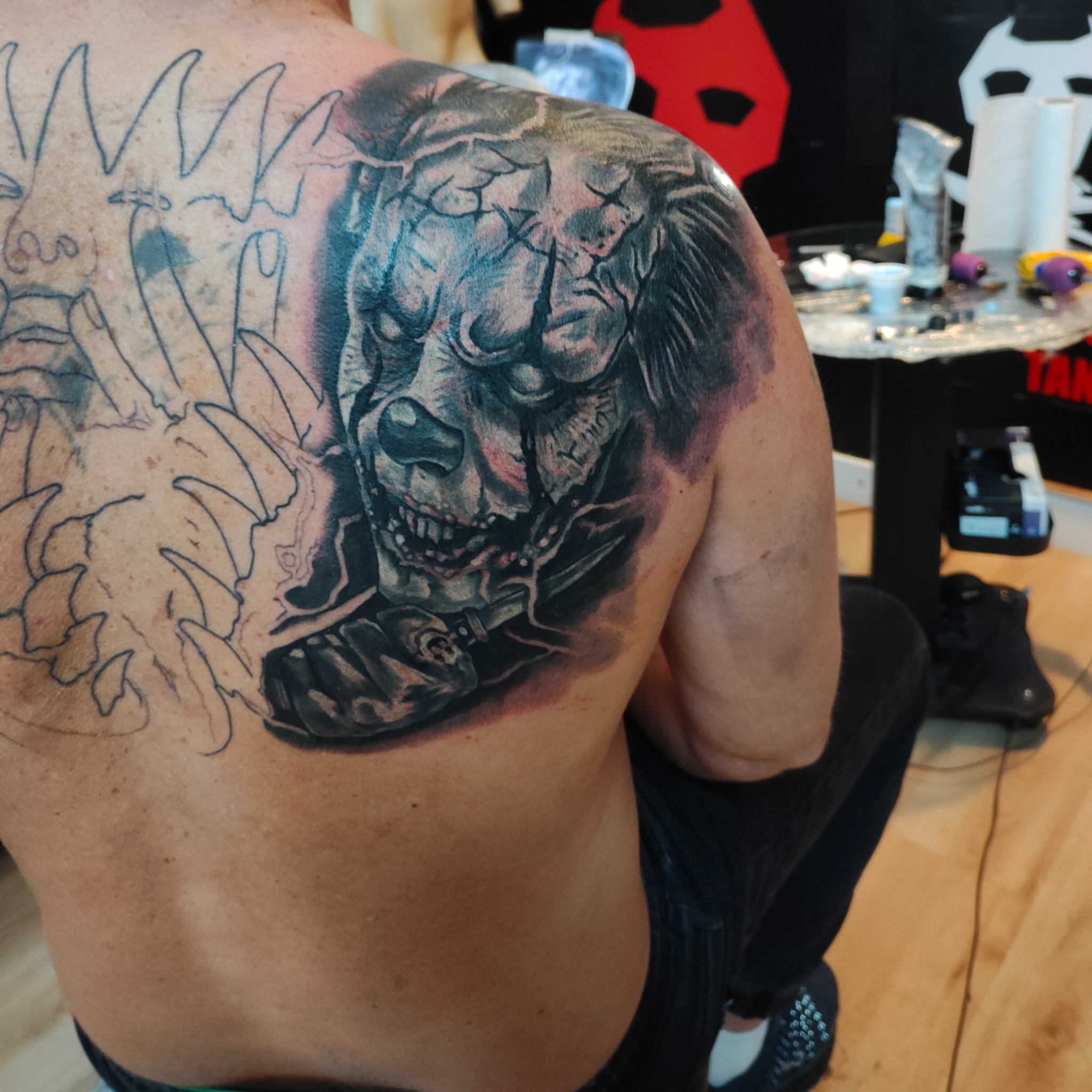 Tatuowanie pleców: mroczny klaun z nożem, obok zarys drugiego tatuażu. Widoczne narzędzia pracy i fragment ubrania osoby tatuowanej.