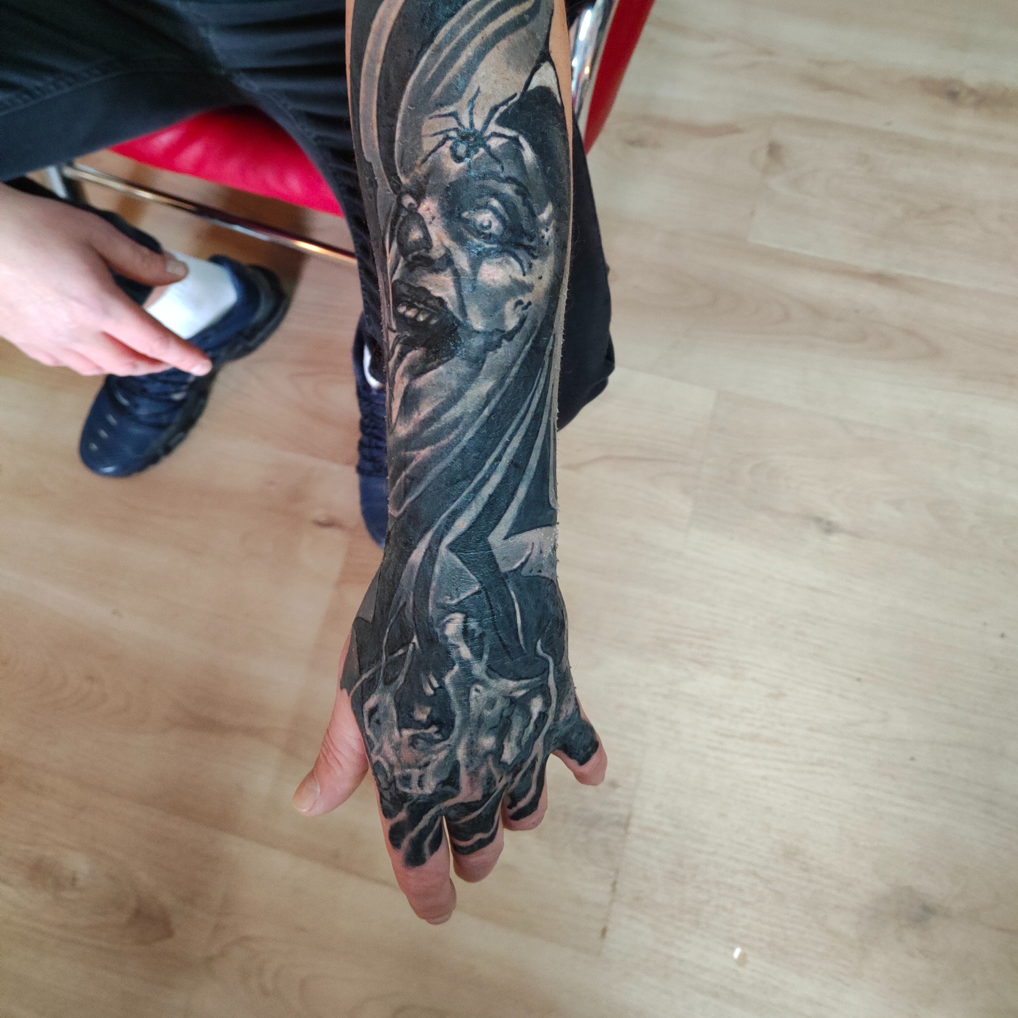 Ręka z czarno-szarym tatuażem w stylu horroru, przedstawiającym twarz z pająkiem. Wykonanie tatuażu w Warszawie, widok z góry na drewnianą podłogę.