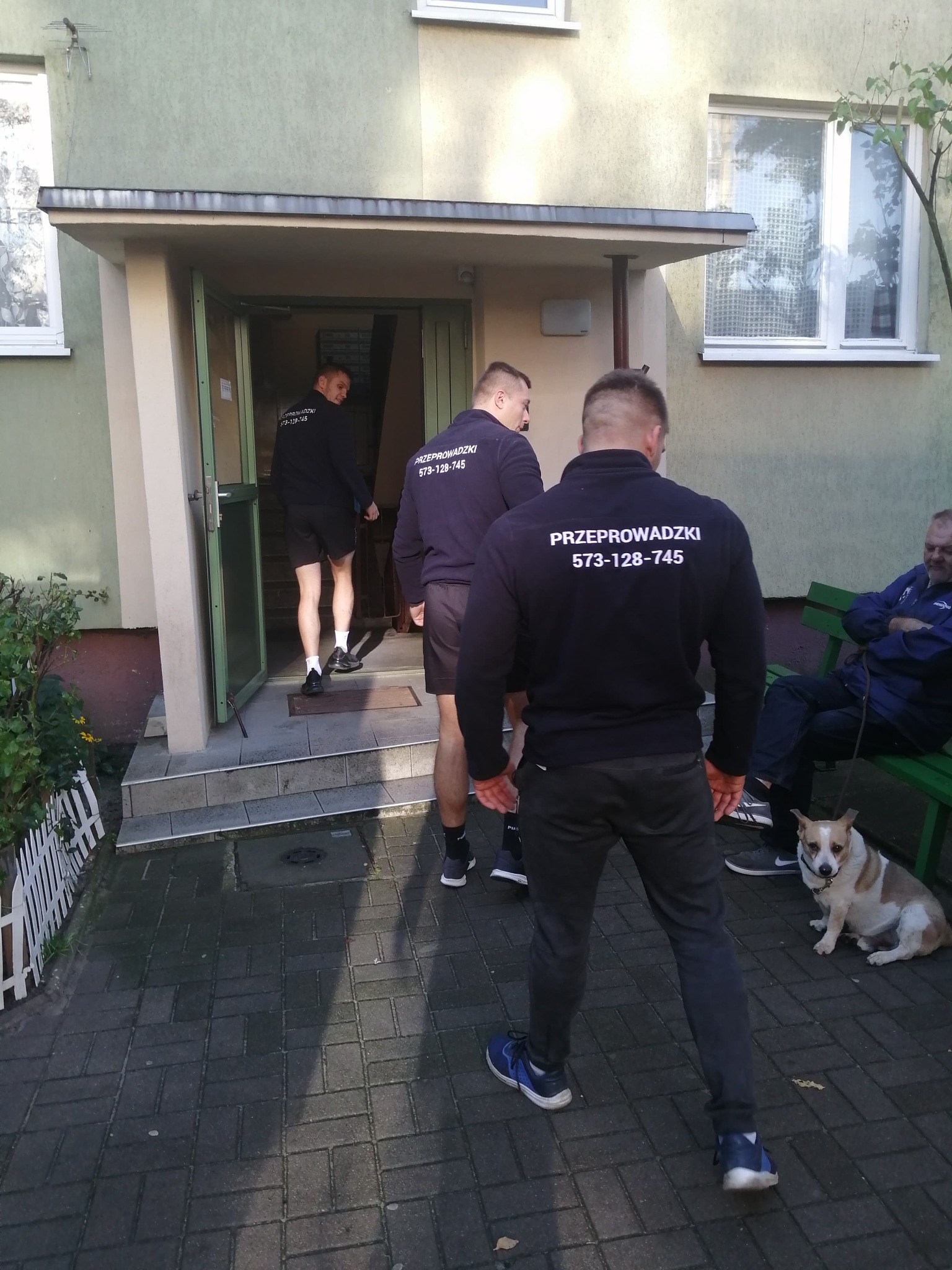 Ekipa w granatowych bluzach z napisem 'Przeprowadzki' wchodzi do budynku; mężczyzna siedzi na ławce obok psa. Widok z zewnątrz.