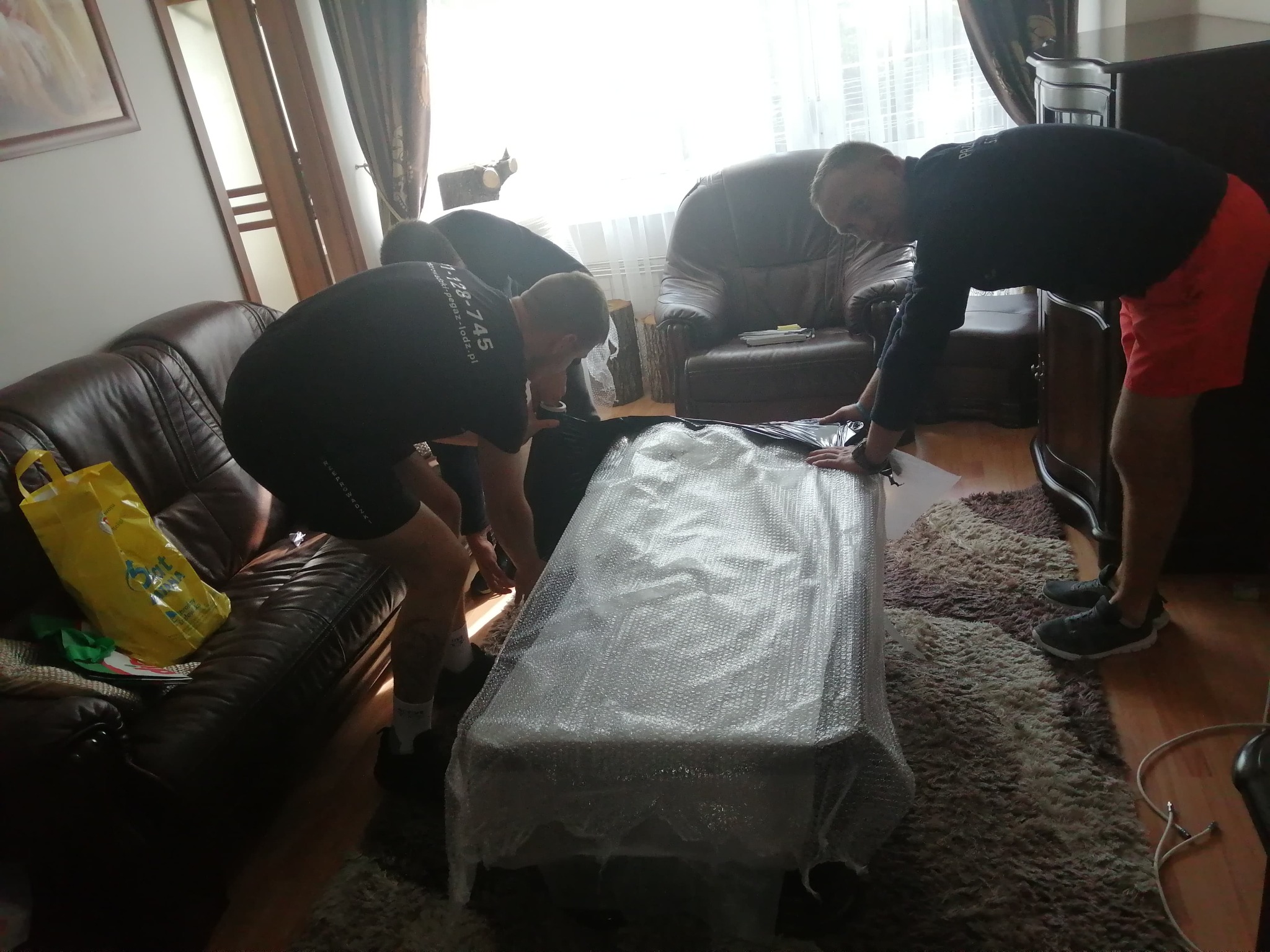 Trzech mężczyzn owija mebel folią bąbelkową w trakcie przeprowadzki. W tle skórzane sofy i dywan. Jeden z mężczyzn ma na koszulce logo firmy.