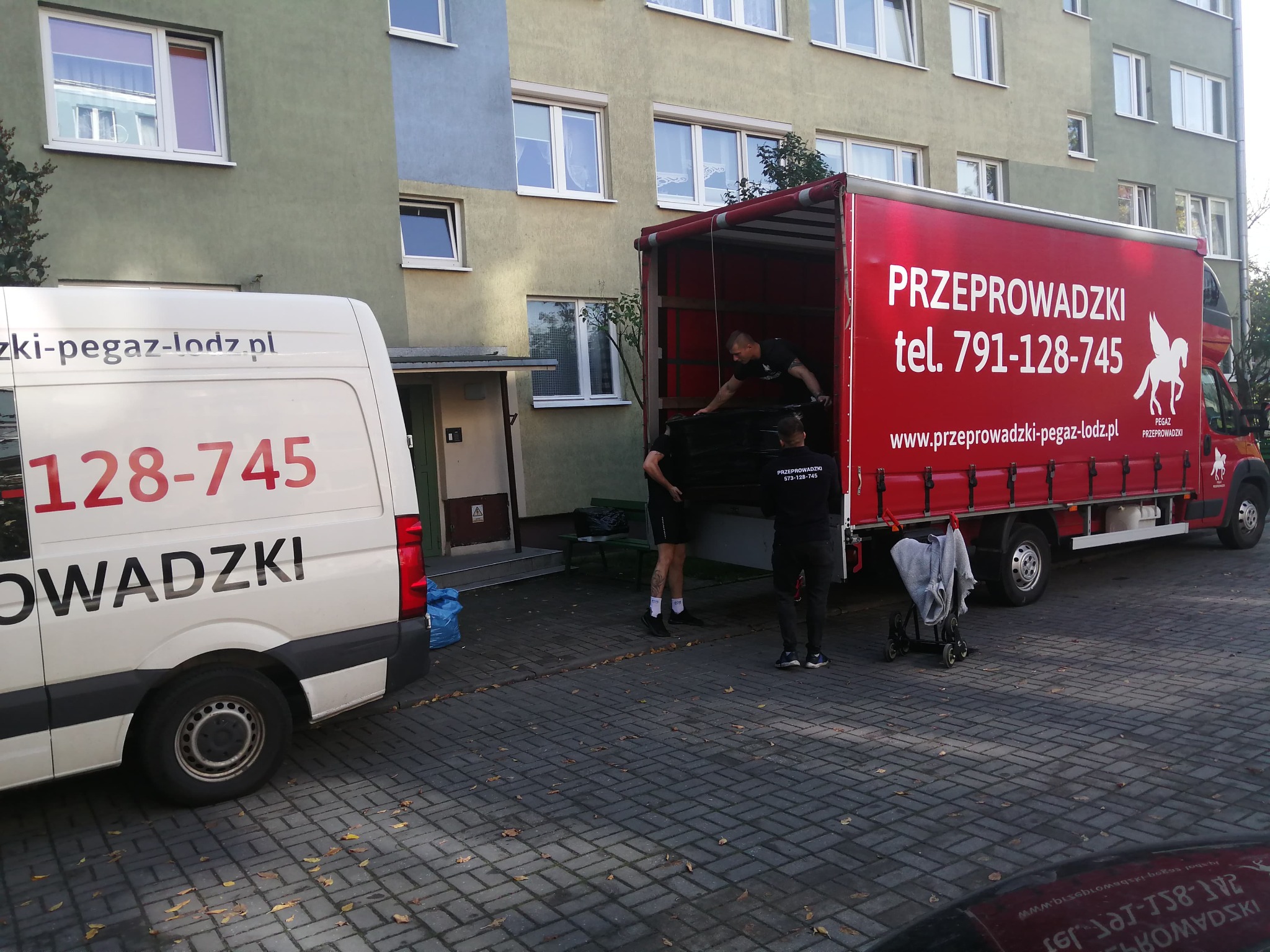 Załadunek mebli do czerwonej ciężarówki z napisem 'Przeprowadzki' na tle bloku mieszkalnego i białego busa z logo firmy. Pracownicy w trakcie przenoszenia.