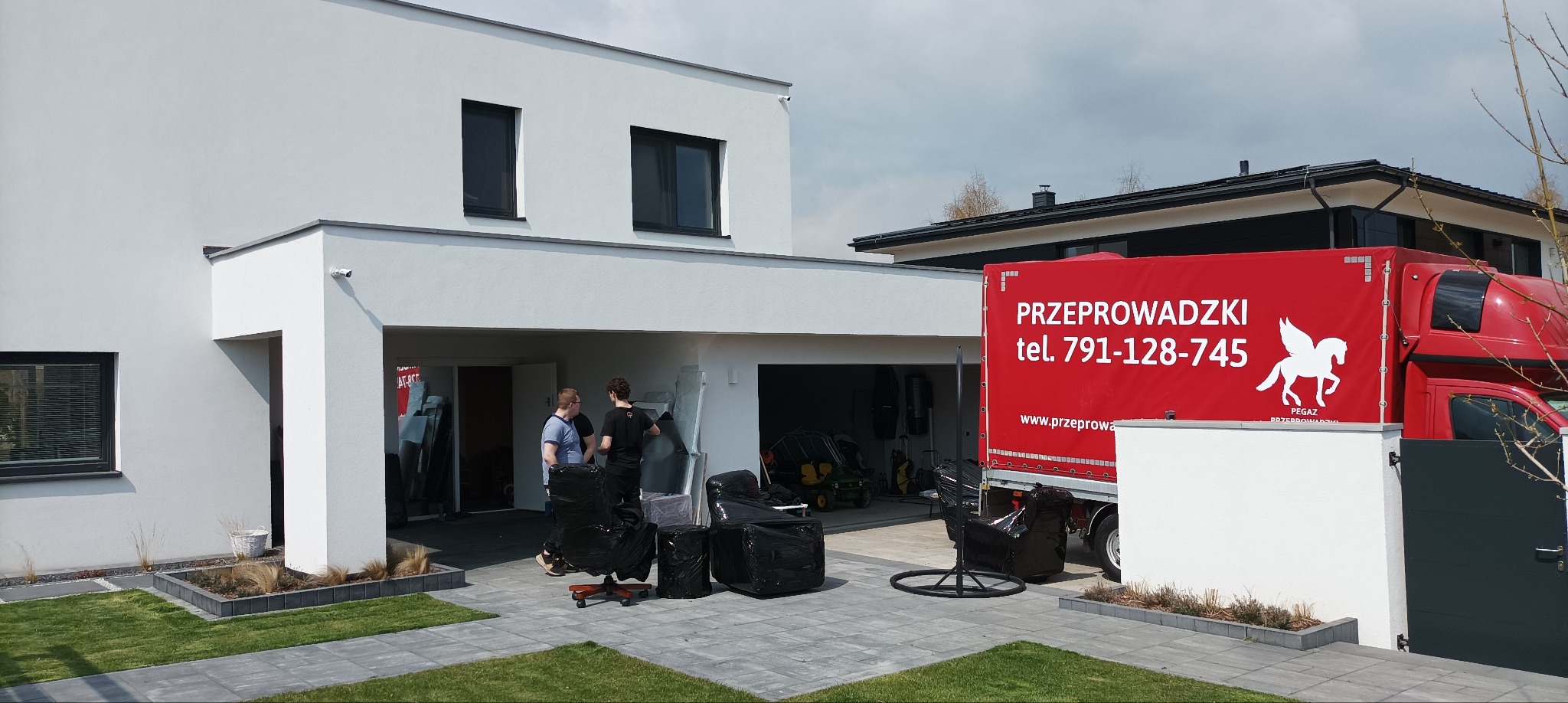 Przeprowadzka z nowoczesnego domu: meble owinięte folią, ekipa i czerwona ciężarówka z logo firmy przeprowadzkowej przed budynkiem.