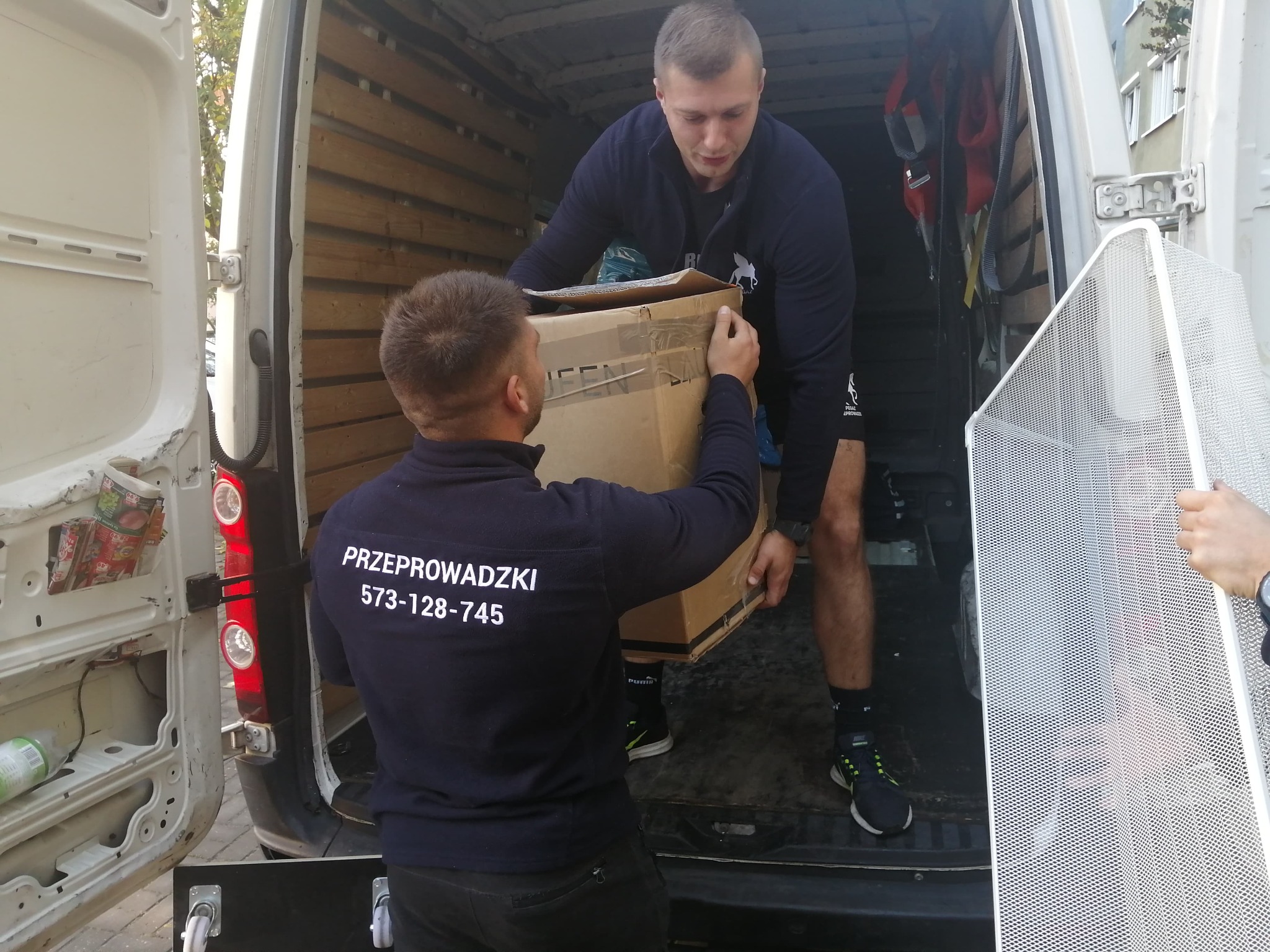Dwóch mężczyzn w trakcie załadunku kartonu do furgonetki. Na plecach jednego z nich widnieje napis 'Przeprowadzki' i numer telefonu.