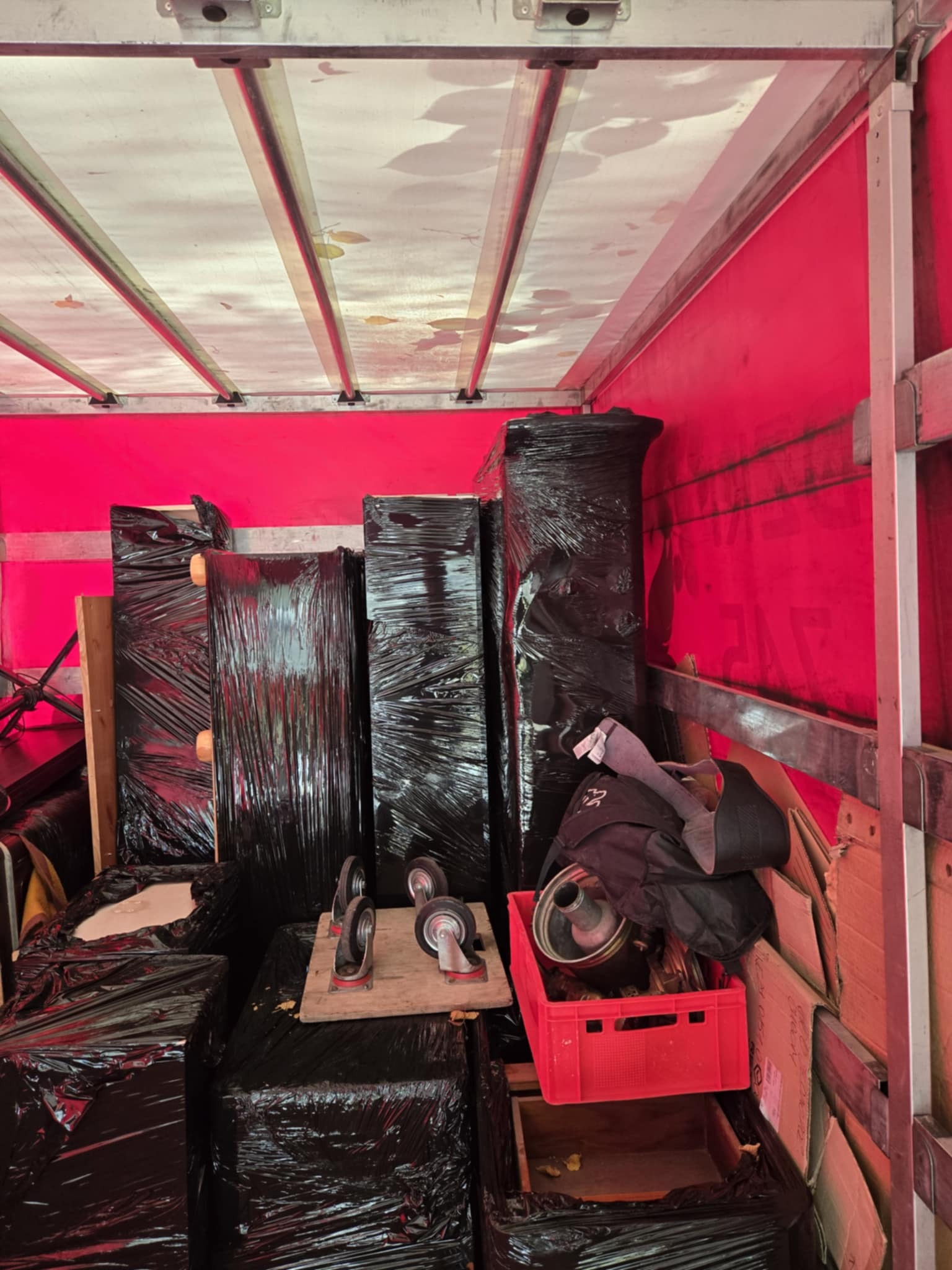 Wnętrze czerwonej furgonetki wypełnionej meblami i przedmiotami zabezpieczonymi czarną folią, gotowymi do transportu. Widoczna skrzynka i wózek transportowy.