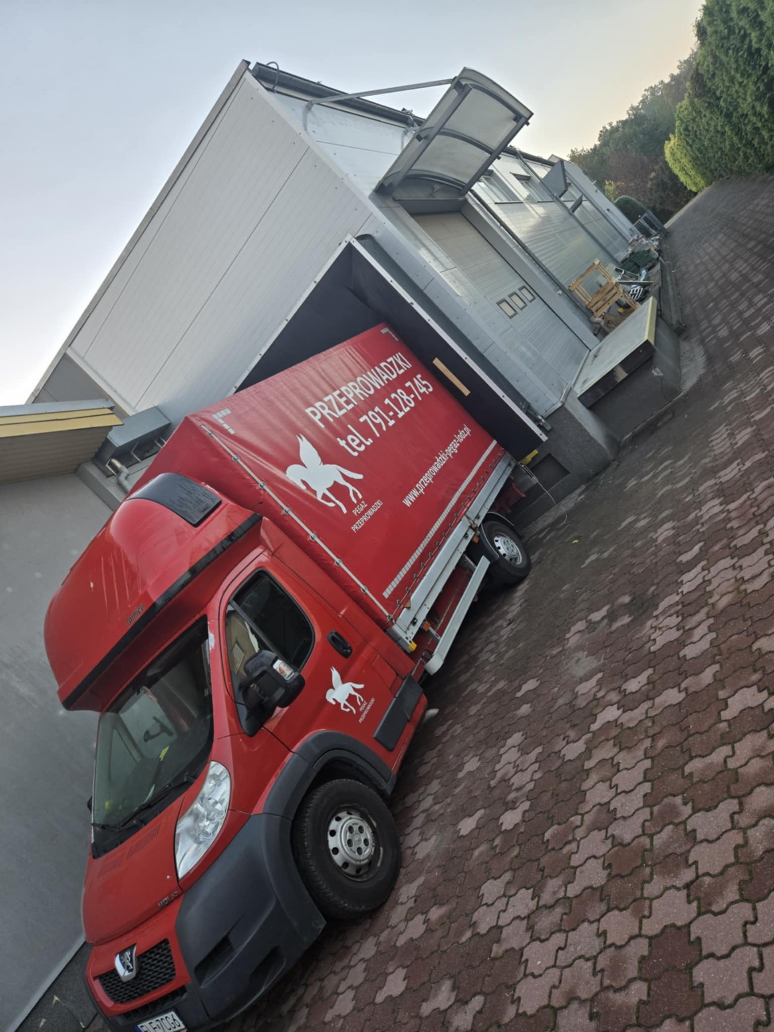 Czerwona ciężarówka firmy przeprowadzkowej Pegaz częściowo w doku załadunkowym, widoczny numer telefonu i adres strony internetowej na plandece.