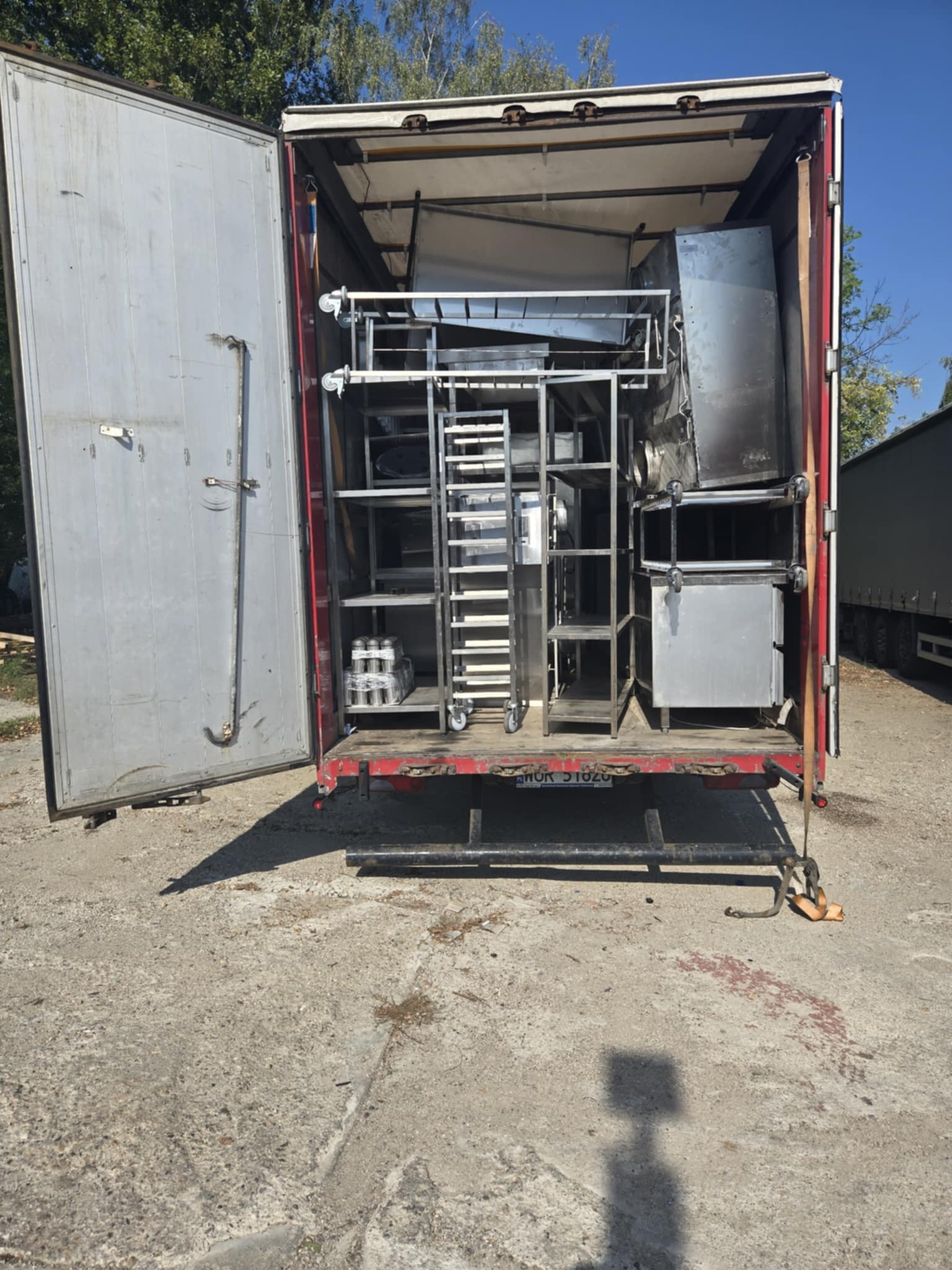 Otwarta naczepa z metalowymi konstrukcjami i urządzeniami na kółkach, gotowymi do transportu. Widoczny jasny niebieski niebo i drzewa w tle.