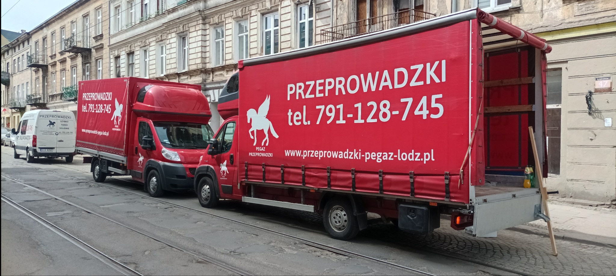 Flota czerwonych samochodów dostawczych z logo Pegaz Przeprowadzki, zaparkowana na ulicy z torami tramwajowymi, gotowa do transportu. Otwarta paka jednego z aut.