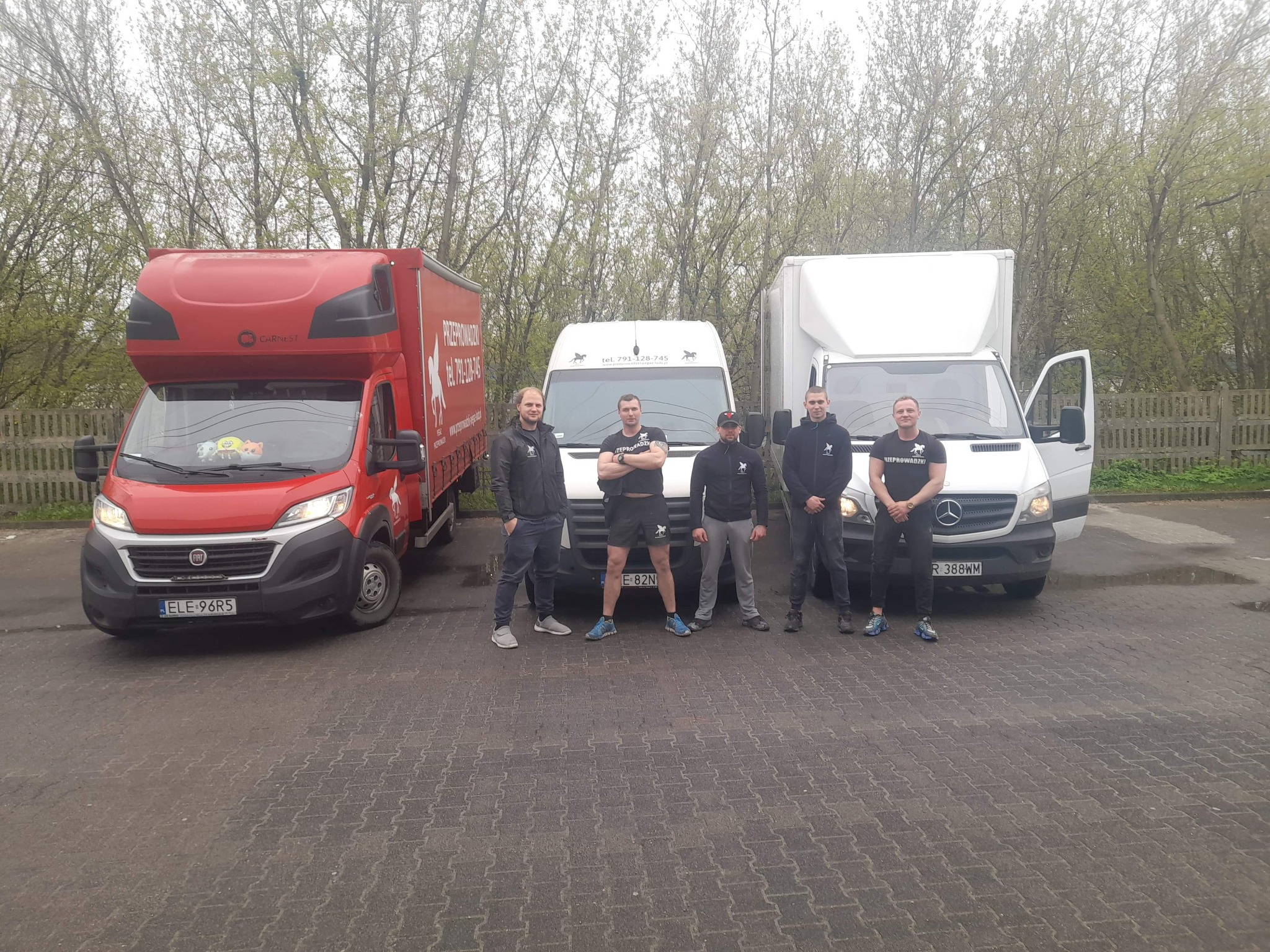 Zespół firmy przeprowadzkowej pozuje przed trzema samochodami dostawczymi z logo firmy na tle drzew. Jeden samochód ma otwarte drzwi.