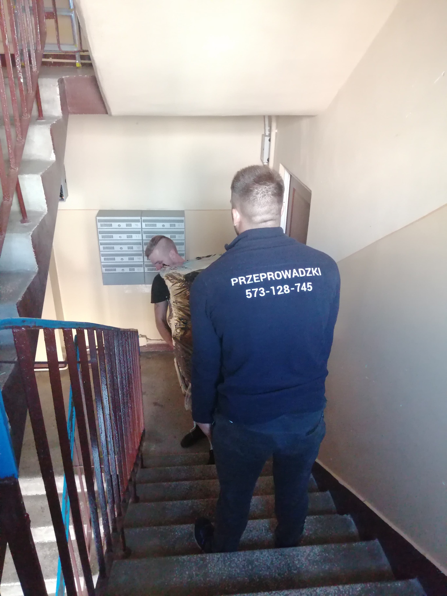 Pracownik firmy przeprowadzkowej w bluzie z numerem telefonu niesie zabezpieczony mebel po schodach w bloku mieszkalnym.