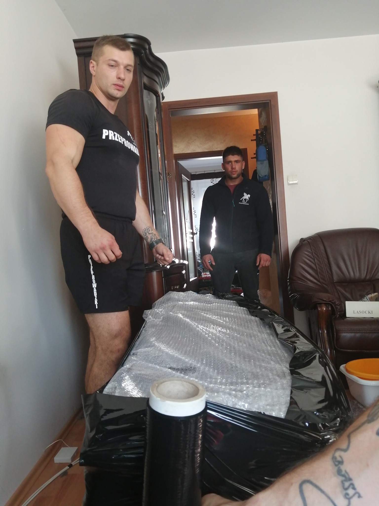 Dwóch mężczyzn w koszulkach z napisem 'Przeprowadzki' zabezpiecza mebel folią bąbelkową i czarną folią stretch w jasnym wnętrzu podczas przeprowadzki.