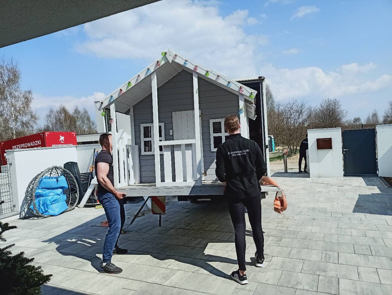 Przenoszenie domku dla dzieci na przyczepie transportowej. Dwóch mężczyzn przenosi konstrukcję na tle błękitnego nieba i zabudowań.