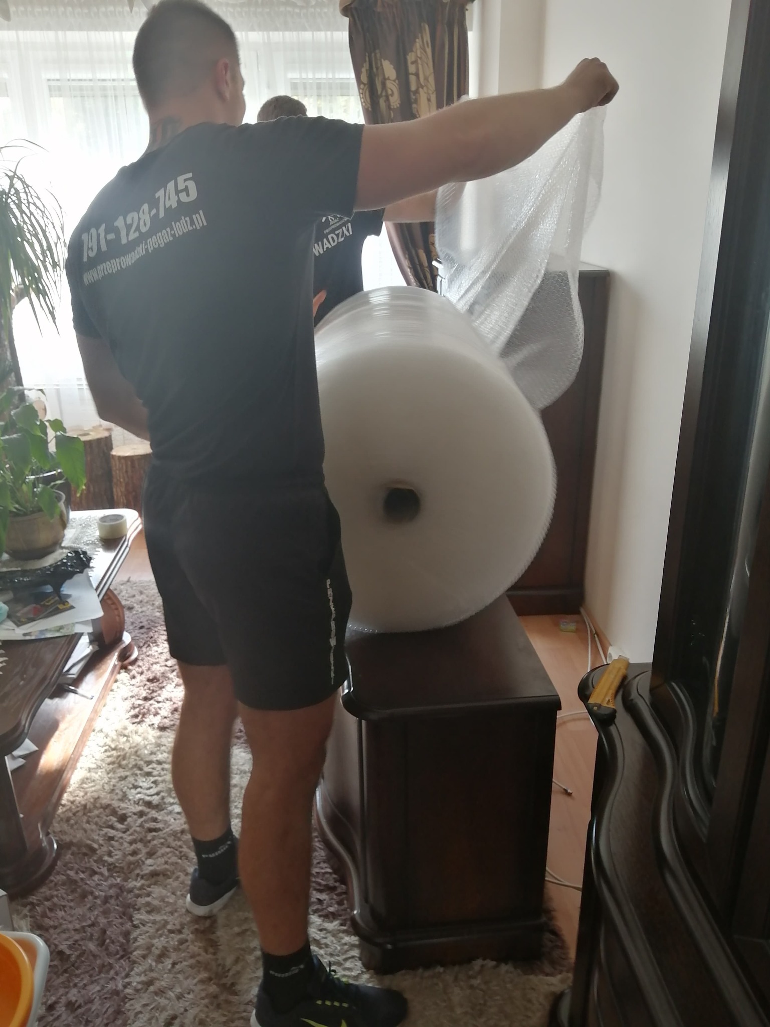 Pracownik firmy przeprowadzkowej owija komodę folią bąbelkową. Widoczny numer telefonu na koszulce pracownika i rolka folii. Przygotowanie do transportu mebli.