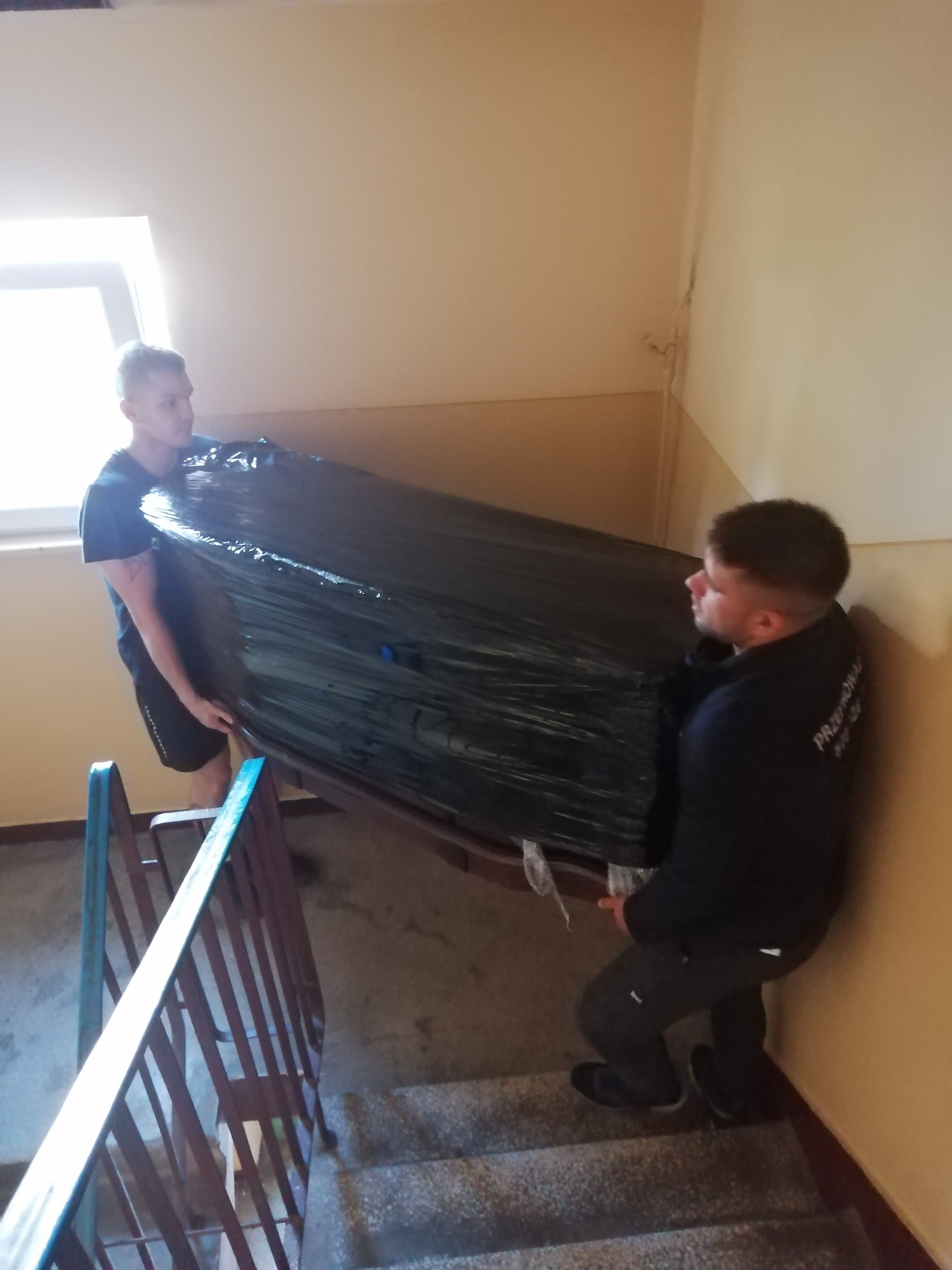 Dwóch mężczyzn wnosi owinięty folią mebel po schodach na klatce schodowej podczas przeprowadzki. Widoczne barierki i okno.