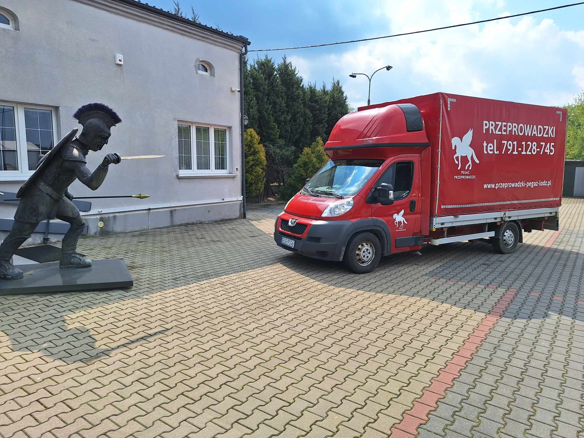 Czerwony samochód dostawczy z logo firmy przeprowadzkowej Pegaz, stojący obok budynku z rzeźbą wojownika. Widoczny numer telefonu i adres strony internetowej.