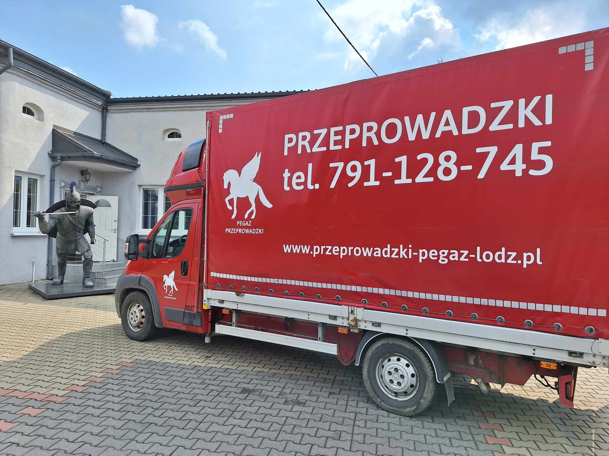 Czerwony samochód dostawczy z reklamą firmy przeprowadzkowej Pegaz, zaparkowany na tle budynku z rzeźbą rycerza. Widoczny numer telefonu i adres strony internetowej.