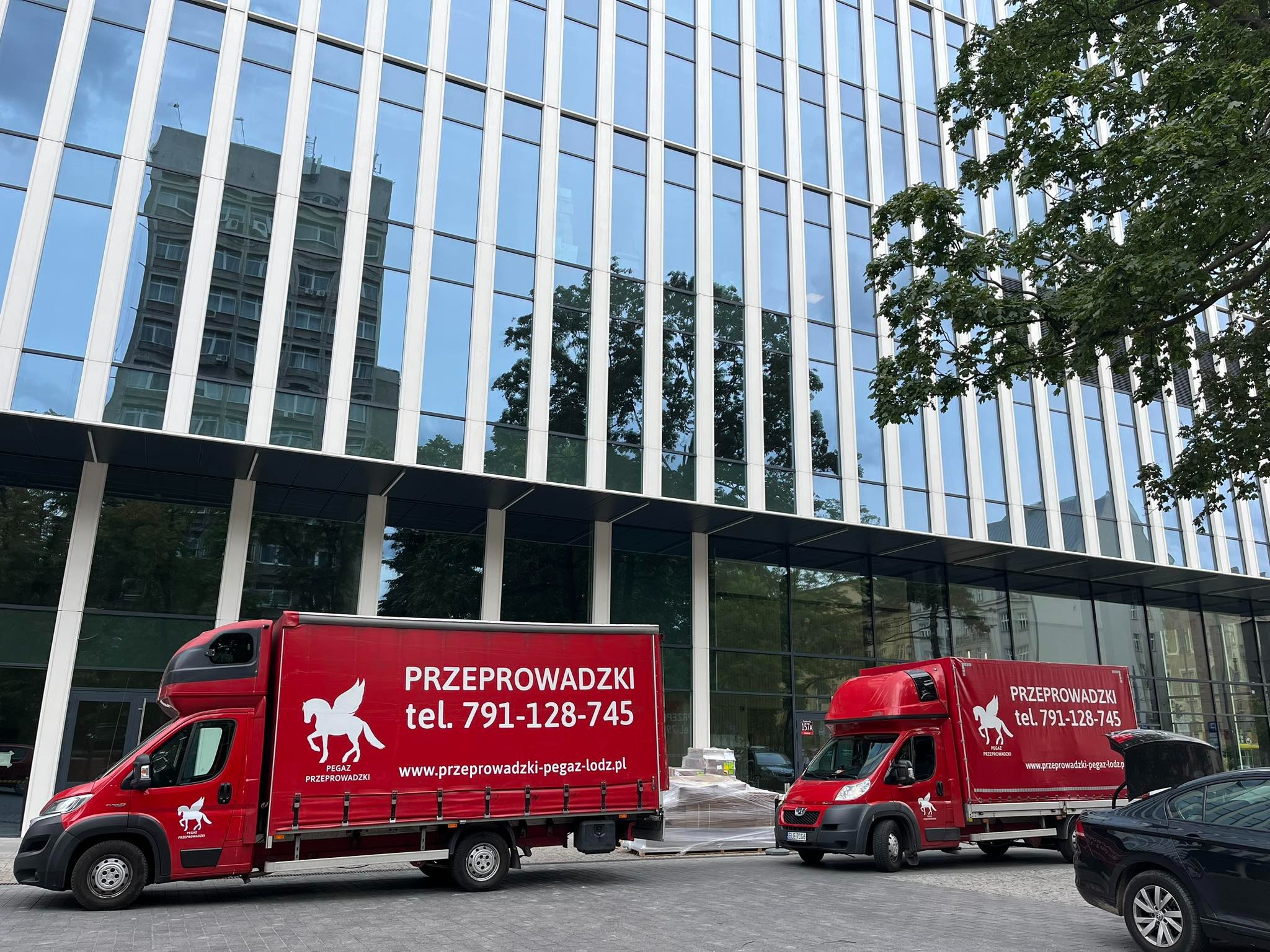 Dwa czerwone busy z logo Pegaz Przeprowadzki na tle nowoczesnego biurowca z lustrzanymi oknami. Jeden bus zaparkowany, drugi w trakcie załadunku.