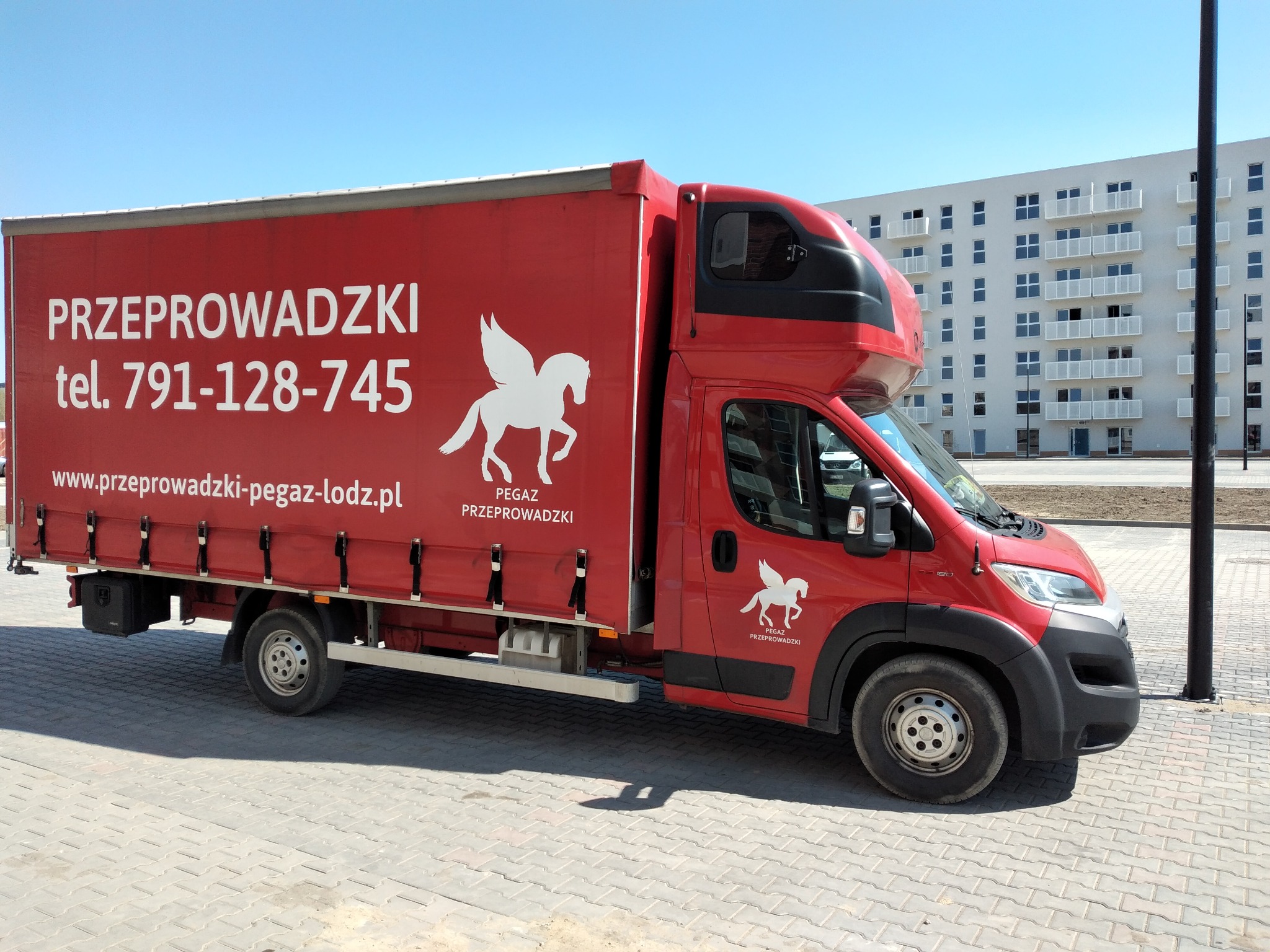 Czerwony samochód dostawczy z logo Pegaz Przeprowadzki na tle jasnego budynku mieszkalnego w słoneczny dzień. Widok z boku, skupienie na detalu graficznym i kolorystyce.