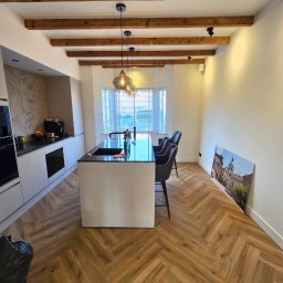 Firma Remontowo-budowlana Patryk Plech - Nowoczesna kuchnia z drewnianą podłogą ułożoną w jodełkę, wyspą z czarnym blatem i drewnianymi belkami na suficie. Na ścianie obraz z architekturą miejską.