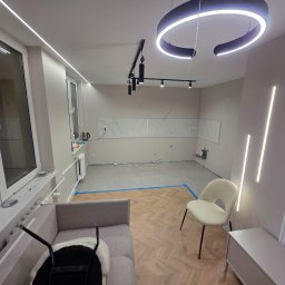 Firma Remontowo-budowlana Patryk Plech - Jasne wnętrze w trakcie wykańczania: parkiet w jodełkę, nowoczesne oświetlenie LED, fragment sofy i krzesła. Widoczne przygotowania do montażu płytek.