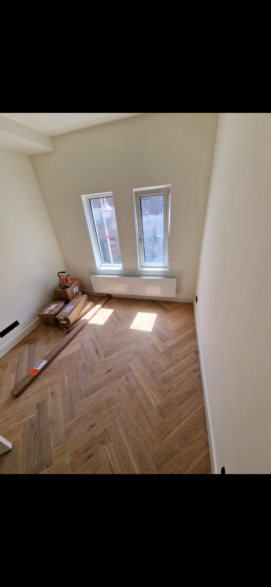 Jasne wnętrze poddasza z ułożonym parkietem w jodełkę. Pod oknami grzejnik, obok paczki z panelami i zwinięty kabel. Świeżo po remoncie, gotowe do umeblowania.