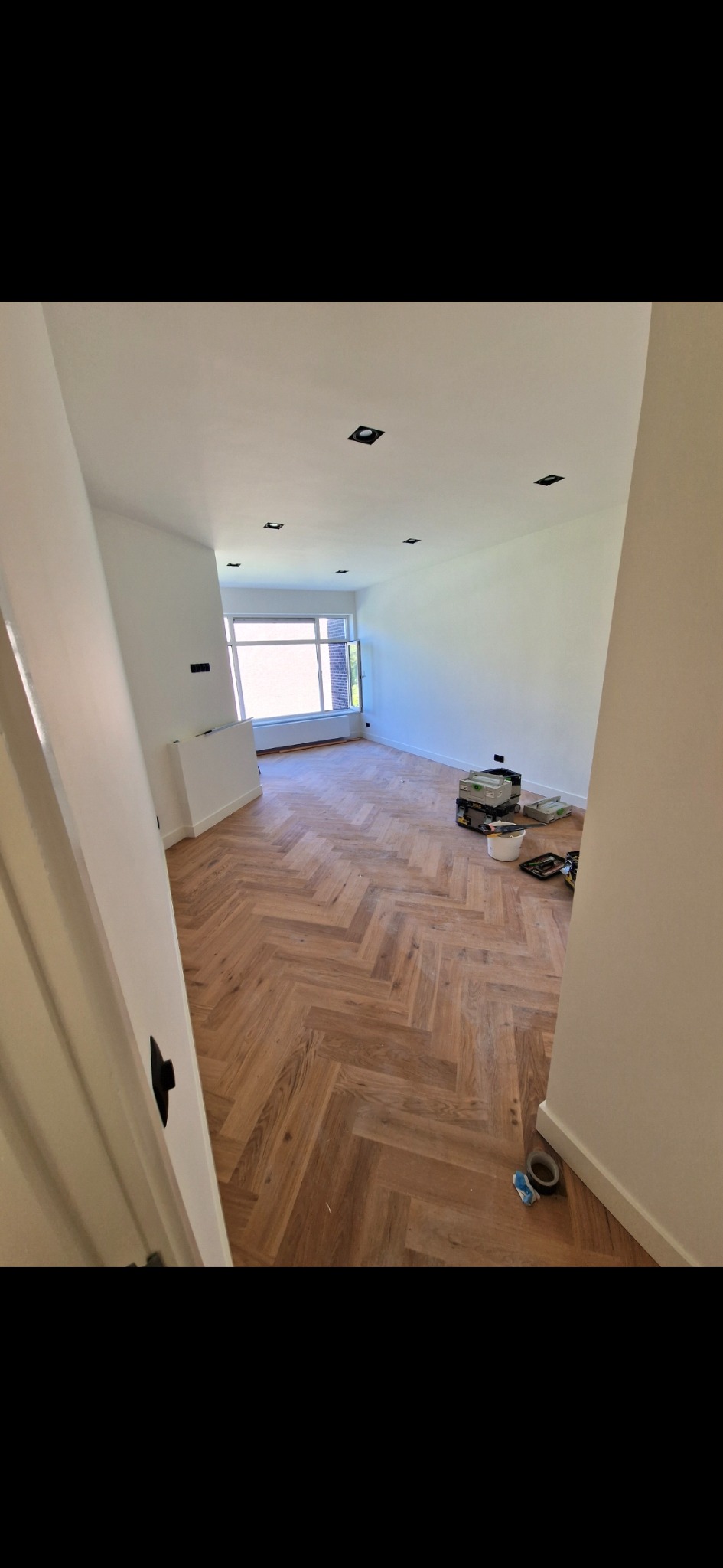 Jasne wnętrze po remoncie z parkietem w jodełkę. Widoczne narzędzia i materiały malarskie. Świeżo pomalowane ściany i sufit z zamontowanym oświetleniem.