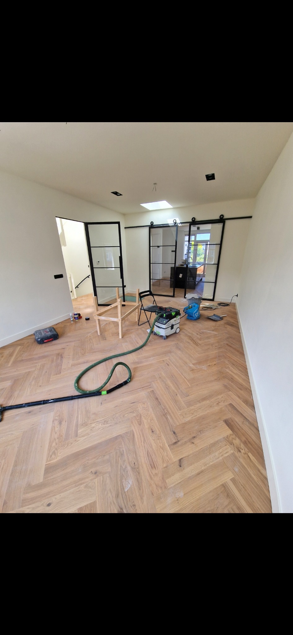 Jasny parkiet w jodełkę po renowacji, widoczne narzędzia i odkurzacz. W tle czarne, przesuwne drzwi w stylu loftowym i białe ściany. Ujęcie z góry, szeroki kadr.