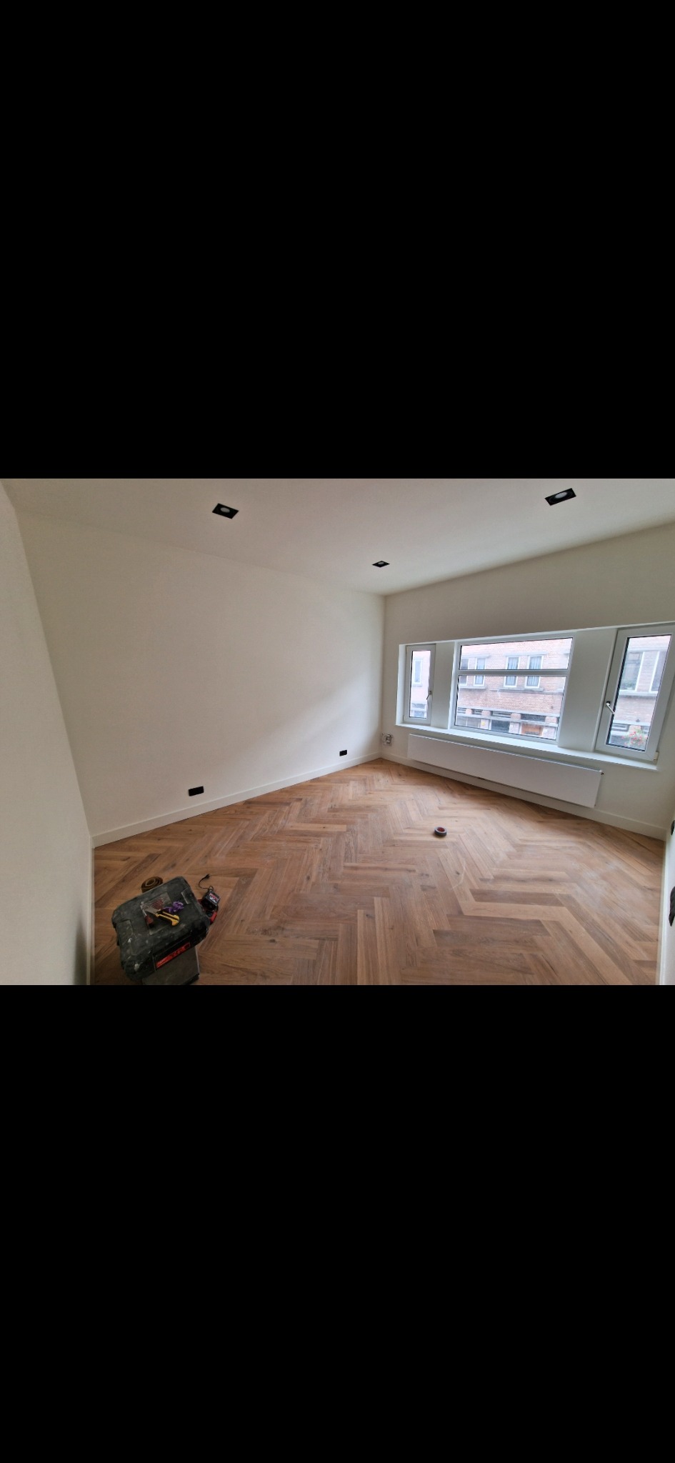 Jasne parkiet w jodełkę francuską w pokoju z białymi ścianami i oknem. Skrzynka z narzędziami w rogu. Widok na budynek z okna.