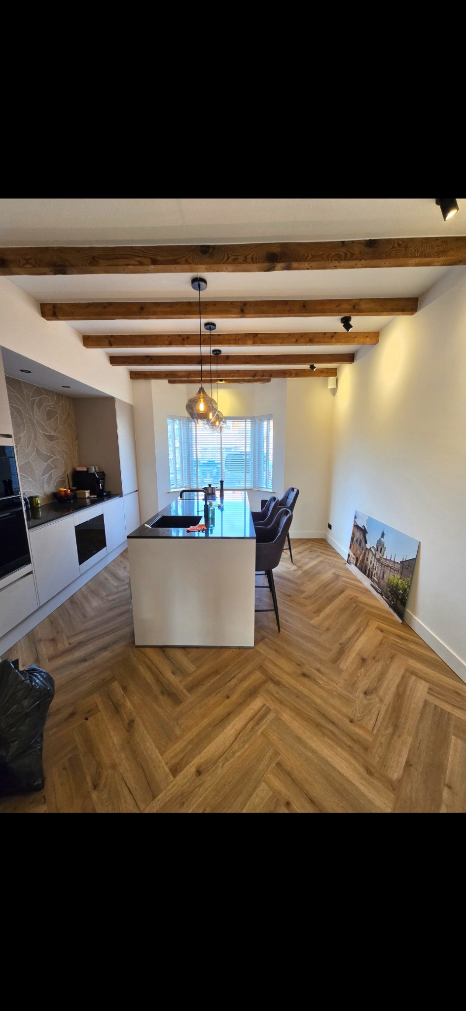 Nowoczesna kuchnia z drewnianą podłogą ułożoną w jodełkę, wyspą z czarnym blatem i drewnianymi belkami na suficie. Na ścianie obraz z architekturą miejską.