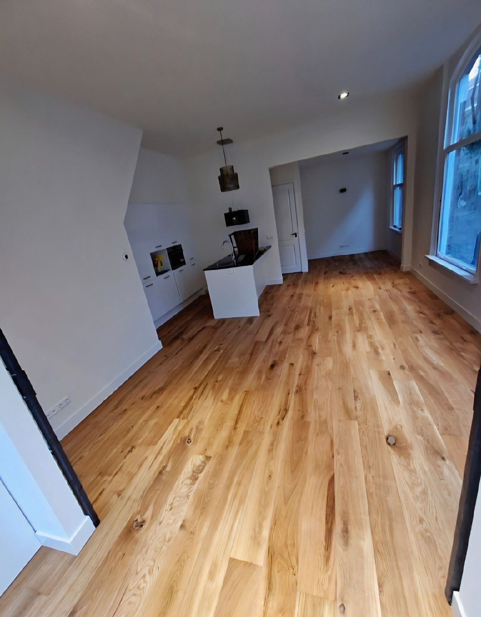 Jasne, drewniane deski podłogowe w nowocześnie wykończonym wnętrzu z białą kuchnią i oknami. Widok na przestronne, odnowione pomieszczenie.