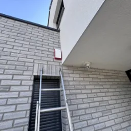 Biała kamera monitoringu i sygnalizator alarmowy na elewacji z cegły. Aluminiowa drabina oparta o ścianę. Widok z dołu na budynek.