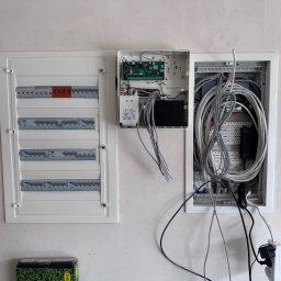 Antoni Mazurkiewicz - Rozdzielnica elektryczna z bezpiecznikami i okablowaniem w białej obudowie na ścianie. Widoczna instalacja alarmowa i zasilacz. Częściowo otwarte skrzynki.
